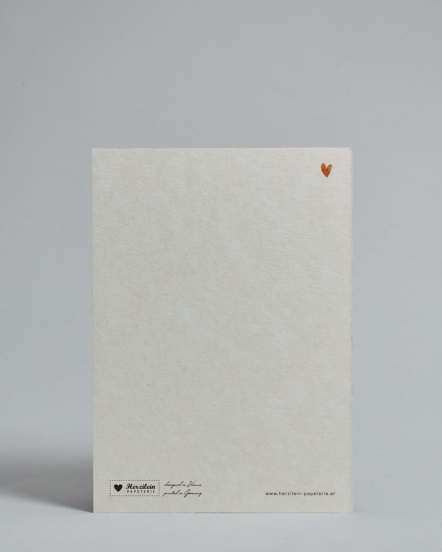 Die Rückseite der Postkarte im Hochformat vor hellgrauem Hintergrund. Rechts oben ist in kleiner Ausführung das gleiche orange Herz wie auf der Vorderseite zu sehen. 
Unten sind klein das Logo Herzilein-Papeterie, der Text: "designed in VIenna, printed in Germany" und "www.herzilein-papeterie.at" zu sehen. 
Der Großteil ist unbedruckt die Struktur der hochwertigen Holzschliffpappe ist gut zu sehen. Hier ist viel Platz zum Schreiben.