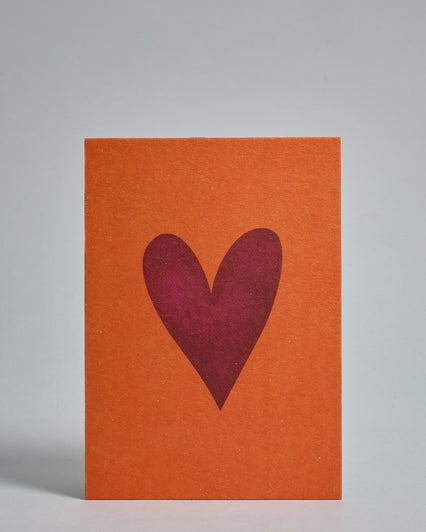 Die Vorderseite der Postkarte im Hochformat vor hellgrauem Hintergrund. Die Postkarte zeigt ein bordeaux-farbenes, gezeichnet aussehendes Herz auf orangem Hintergrund, gedruckt auf Holzschliffpappe.