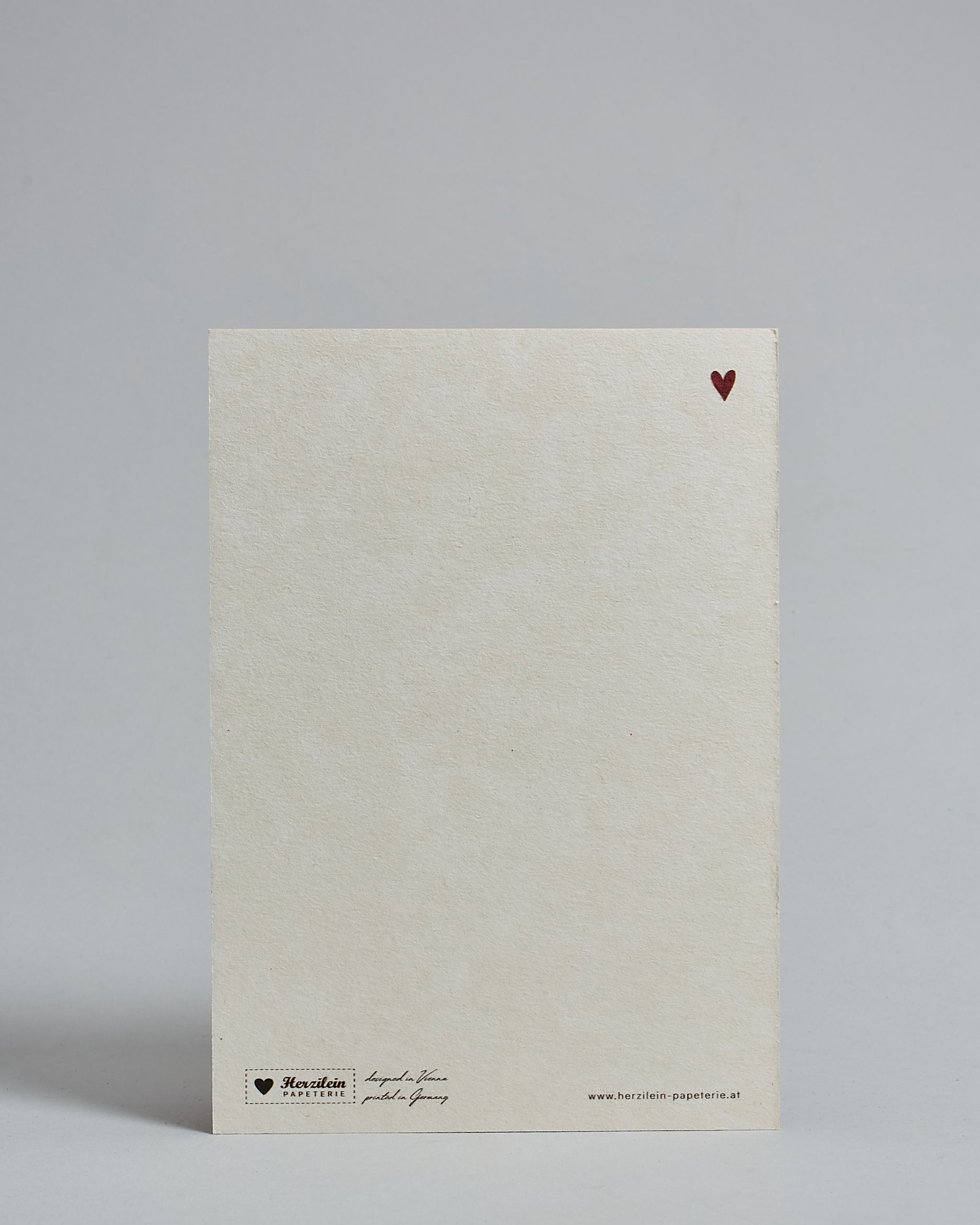 Die Rückseite der Postkarte im Hochformat vor hellgrauem Hintergrund. Rechts oben ist in kleiner Ausführung das gleiche rote Herz wie auf der Vorderseite zu sehen. 
Unten sind klein das Logo Herzilein-Papeterie, der Text: "designed in VIenna, printed in Germany" und "www.herzilein-papeterie.at" zu sehen. 
Der Großteil ist unbedruckt die Struktur der hochwertigen Holzschliffpappe ist gut zu sehen. Hier ist viel Platz zum Schreiben.