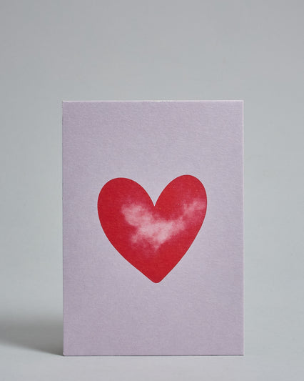 Die Vorderseite der Postkarte im Hochformat vor hellgrauem Hintergrund. Die Postkarte zeigt ein rotes, gezeichnet aussehendes Herz auf blasslila Hintergrund, gedruckt auf Holzschliffpappe.