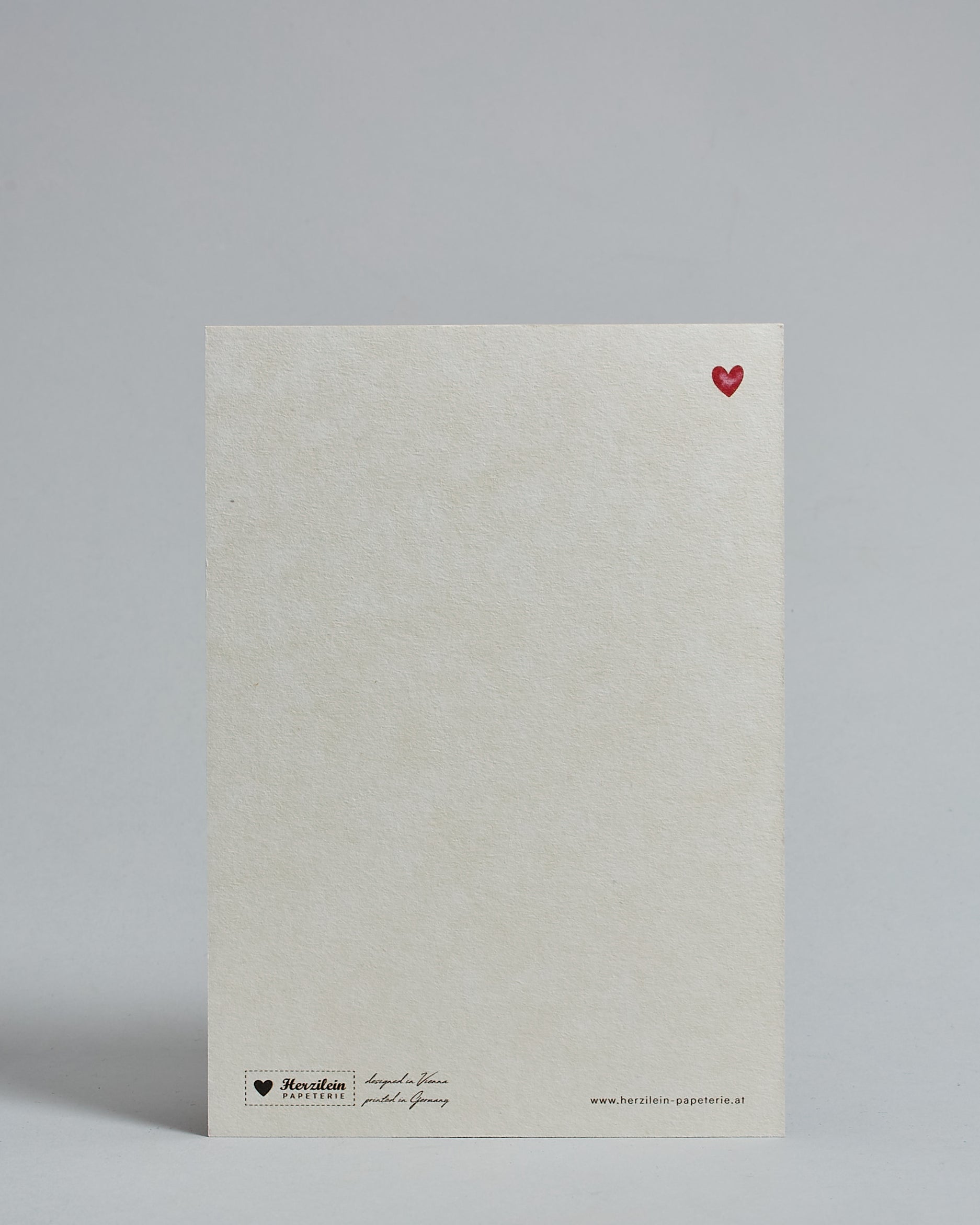 Die Rückseite der Postkarte im Hochformat vor hellgrauem Hintergrund. Rechts oben ist in kleiner Ausführung das gleiche rote Herz wie auf der Vorderseite zu sehen. 
Unten sind klein das Logo Herzilein-Papeterie, der Text: "designed in VIenna, printed in Germany" und "www.herzilein-papeterie.at" zu sehen. 
Der Großteil ist unbedruckt die Struktur der hochwertigen Holzschliffpappe ist gut zu sehen. Hier ist viel Platz zum Schreiben