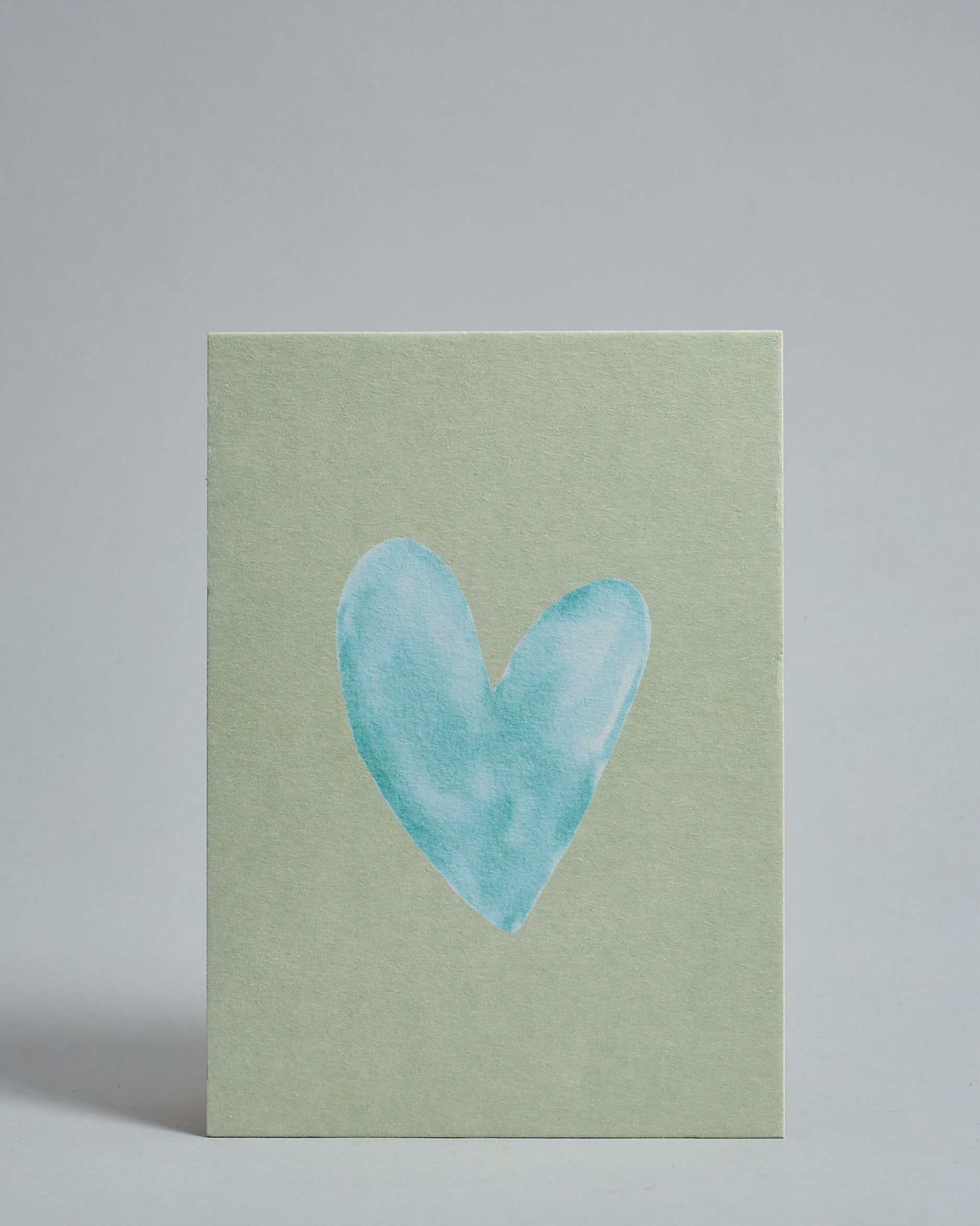 Die Vorderseite der Postkarte im Hochformat vor hellgrauem Hintergrund. Die Postkarte zeigt ein hellblaues, gezeichnet aussehendes Herz auf mint-farbenem Hintergrund, gedruckt auf Holzschliffpappe.