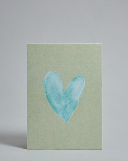 Die Vorderseite der Postkarte im Hochformat vor hellgrauem Hintergrund. Die Postkarte zeigt ein hellblaues, gezeichnet aussehendes Herz auf mint-farbenem Hintergrund, gedruckt auf Holzschliffpappe.
