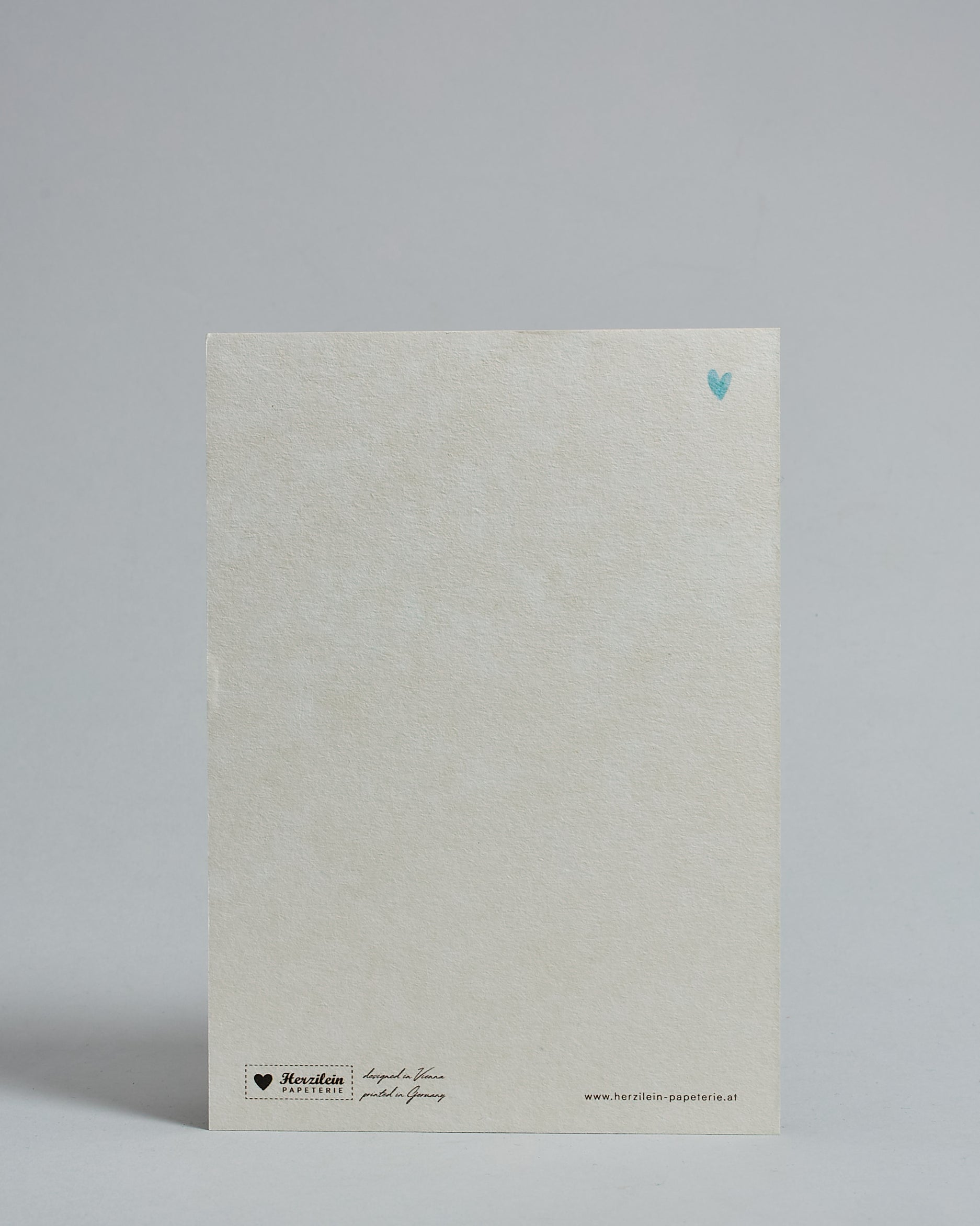 Die Rückseite der Postkarte im Hochformat vor hellgrauem Hintergrund. Rechts oben ist in kleiner Ausführung das gleiche hellblaue Herz wie auf der Vorderseite zu sehen. 
Unten sind klein das Logo Herzilein-Papeterie, der Text: "designed in VIenna, printed in Germany" und "www.herzilein-papeterie.at" zu sehen. 
Der Großteil ist unbedruckt die Struktur der hochwertigen Holzschliffpappe ist gut zu sehen. Hier ist viel Platz zum Schreiben.