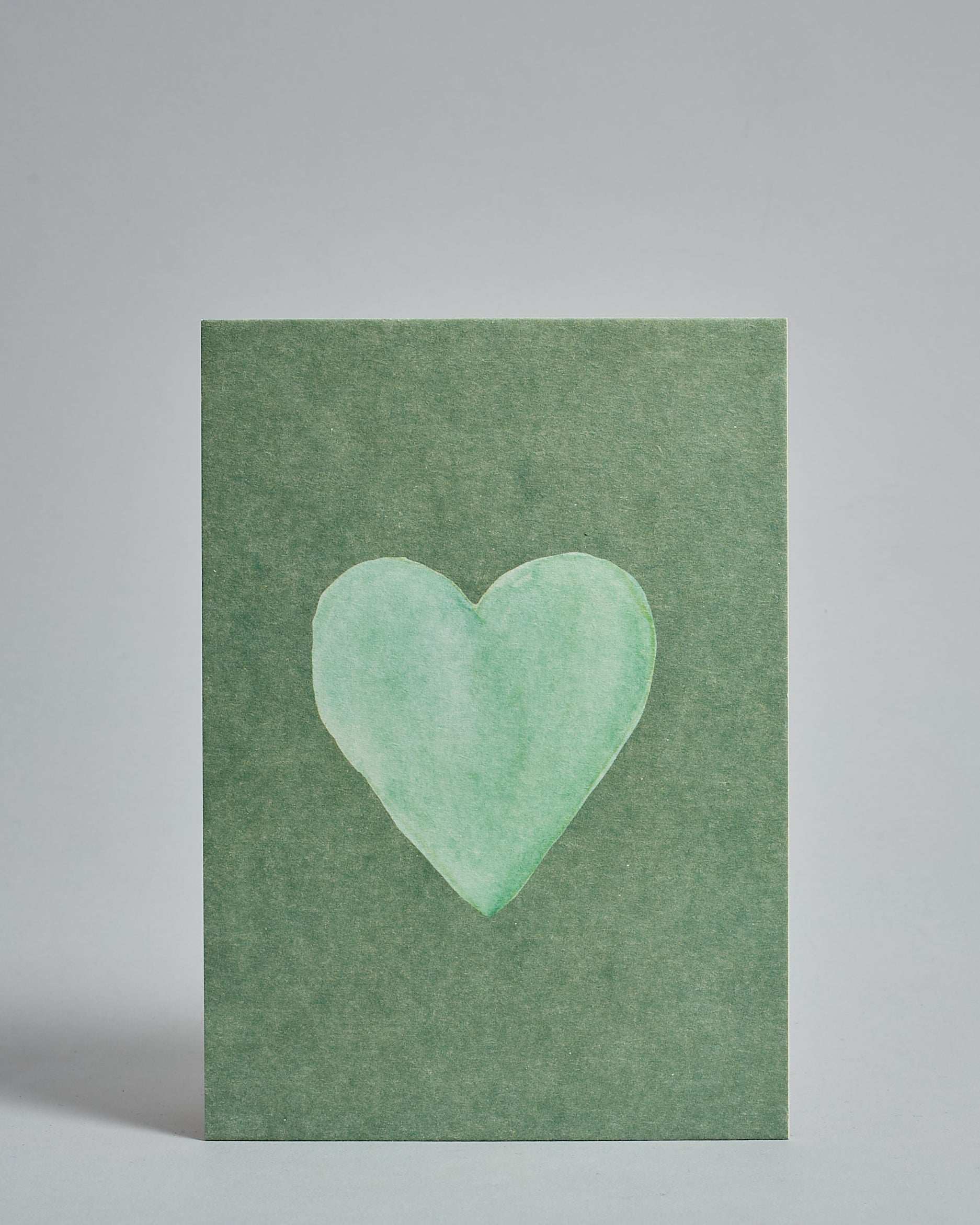 Die Vorderseite der Postkarte im Hochformat vor hellgrauem Hintergrund. Die Postkarte zeigt ein mintfarbenes, gezeichnet aussehendes Herz auf grünem Hintergrund gedruckt auf Holzschliffpappe.