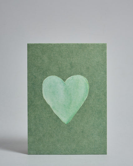 Die Vorderseite der Postkarte im Hochformat vor hellgrauem Hintergrund. Die Postkarte zeigt ein mintfarbenes, gezeichnet aussehendes Herz auf grünem Hintergrund gedruckt auf Holzschliffpappe.