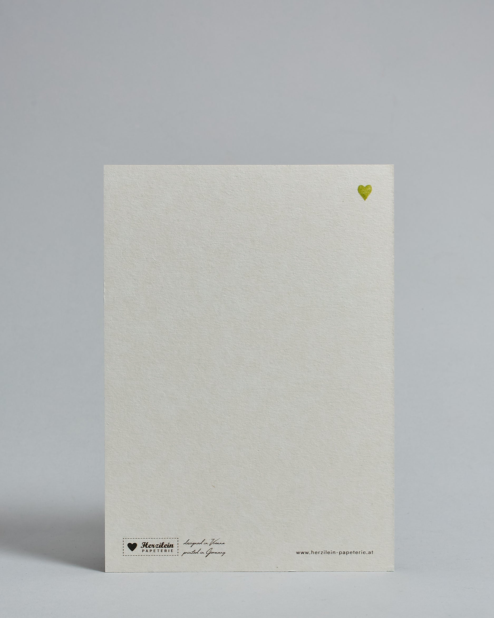Die Rückseite der Postkarte im Hochformat vor hellgrauem Hintergrund. Rechts oben ist in kleiner Ausführung das gleiche grüne Herz wie auf der Vorderseite zu sehen. 
Unten sind klein das Logo Herzilein-Papeterie, der Text: "designed in VIenna, printed in Germany" und "www.herzilein-papeterie.at" zu sehen. 
Der Großteil ist unbedruckt die Struktur der hochwertigen Holzschliffpappe ist gut zu sehen. Hier ist viel Platz zum Schreiben.