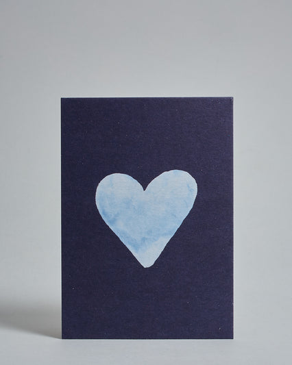 Die Vorderseite der Postkarte im Hochformat vor hellgrauem Hintergrund. Die Postkarte zeigt ein hellblaues, gezeichnet aussehendes Herz auf dunkelblauem Hintergrund, gedruckt auf Holzschliffpappe.