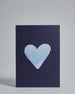 Die Vorderseite der Postkarte im Hochformat vor hellgrauem Hintergrund. Die Postkarte zeigt ein hellblaues, gezeichnet aussehendes Herz auf dunkelblauem Hintergrund, gedruckt auf Holzschliffpappe.