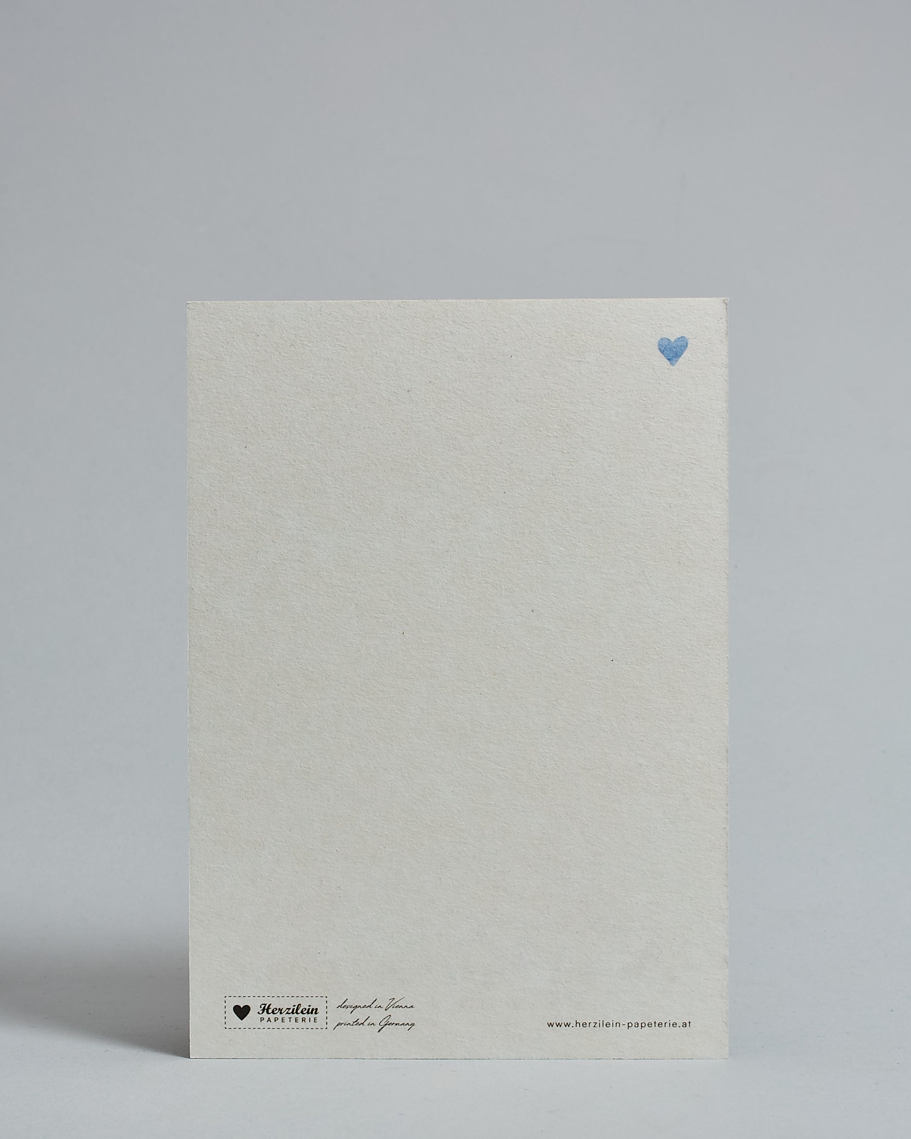 Die Rückseite der Postkarte im Hochformat vor hellgrauem Hintergrund. Rechts oben ist in kleiner Ausführung das gleiche hellblaue Herz wie auf der Vorderseite zu sehen. 
Unten sind klein das Logo Herzilein-Papeterie, der Text: "designed in VIenna, printed in Germany" und "www.herzilein-papeterie.at" zu sehen. 
Der Großteil ist unbedruckt die Struktur der hochwertigen Holzschliffpappe ist gut zu sehen. Hier ist viel Platz zum Schreiben.