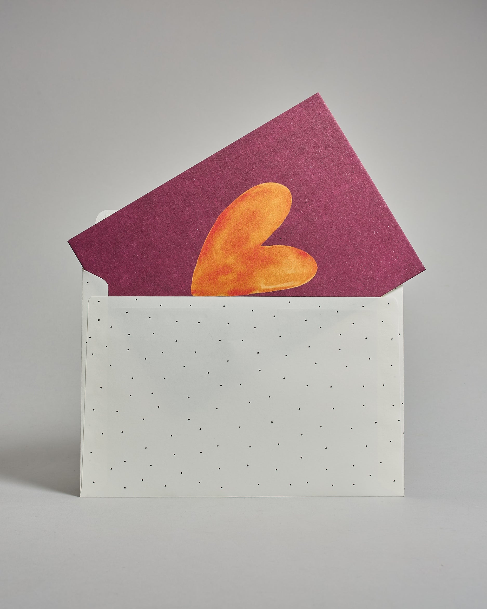 Die Rückseite des weißen Kuverts mit schwarzen Punkten in das schräg eine Postkarte steckt die großteils noch sichtbar ist. Auf der Postkarte ist eine oranges Herz auf dunkelrotem Hinergrund. Das Kuvert eignet sich perfekt für die Postkarten dieser Serie.