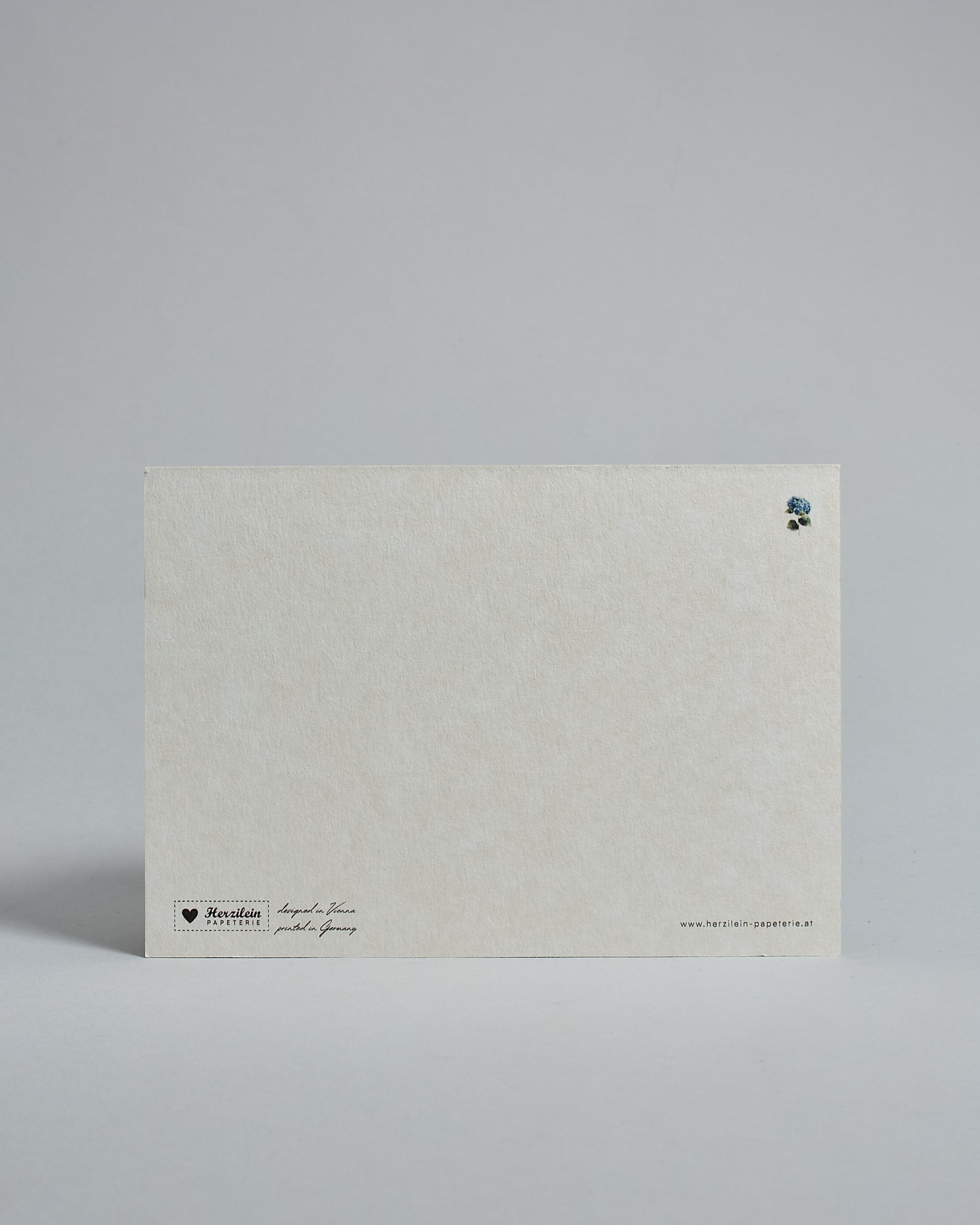 Die Rückseite der Postkarte im Querformat vor hellgrauem Hintergrund. Rechts oben ist in kleiner Ausführung die gleiche Hortensie wie auf der Vorderseite zu sehen. 
Unten sind klein das Logo Herzilein-Papeterie, der Text: "designed in VIenna, printed in Germany" und "www.herzilein-papeterie.at" zu sehen. 
Der Großteil ist unbedruckt die Struktur der hochwertigen Holzschliffpappe ist gut zu sehen. Hier ist viel Platz zum Schreiben.