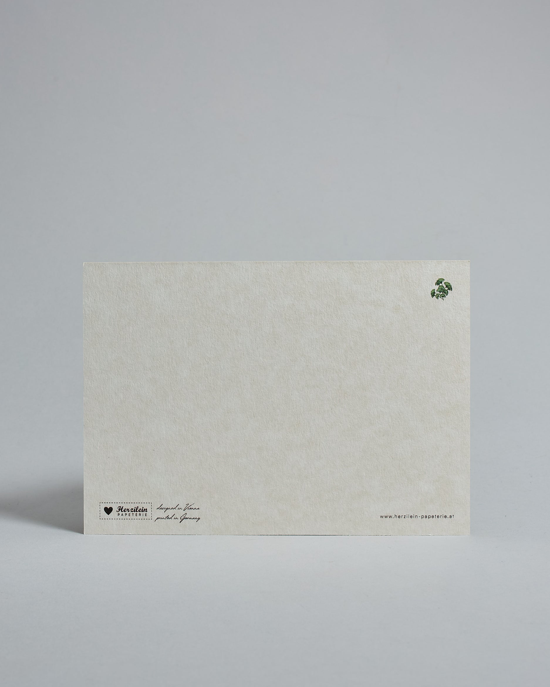 Die Rückseite der Postkarte im Querformat vor hellgrauem Hintergrund. Rechts oben ist in kleiner Ausführung der gleiche Ginko wie auf der Vorderseite zu sehen. 
Unten sind klein das Logo Herzilein-Papeterie, der Text: "designed in VIenna, printed in Germany" und "www.herzilein-papeterie.at" zu sehen. 
Der Großteil ist unbedruckt die Struktur der hochwertigen Holzschliffpappe ist gut zu sehen. Hier ist viel Platz zum Schreiben.