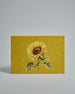 Die Vorderseite der Postkarte im Querformat vor hellgrauem Hintergrund. Die Postkarte zeigt eine gemalte Sonnenblume auf gelbem Hintergrund, gedruckt auf Holzschliffpappe.
