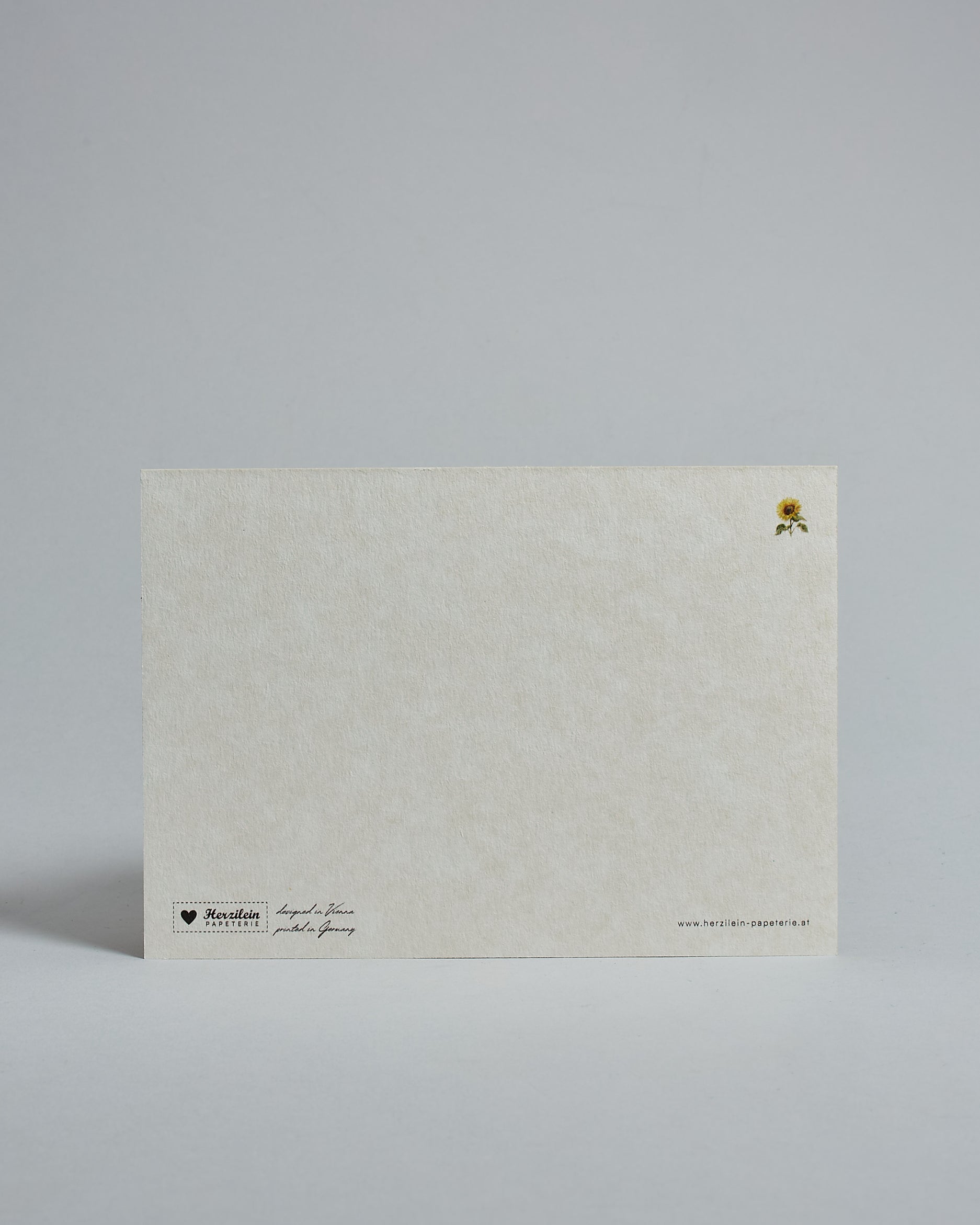 Die Rückseite der Postkarte im Querformat vor hellgrauem Hintergrund. Rechts oben ist in kleiner Ausführung die gleiche Sonnenblume wie auf der Vorderseite zu sehen. 
Unten sind klein das Logo Herzilein-Papeterie, der Text: "designed in VIenna, printed in Germany" und "www.herzilein-papeterie.at" zu sehen. 
Der Großteil ist unbedruckt die Struktur der hochwertigen Holzschliffpappe ist gut zu sehen. Hier ist viel Platz zum Schreiben.