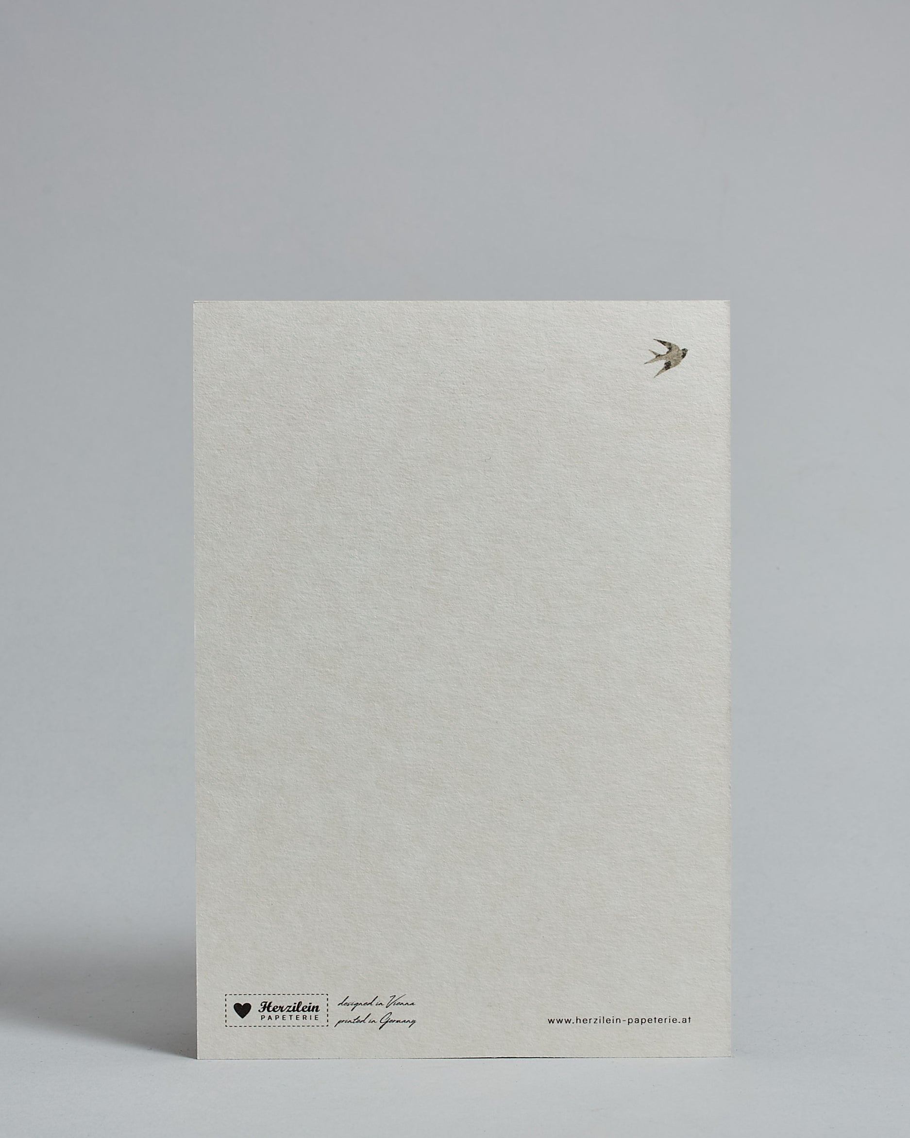 Die Rückseite der Postkarte im Hochformat vor hellgrauem Hintergrund. Rechts oben ist, in kleiner Ausführung, ein Vogel zu sehen. 
Unten sind klein das Logo Herzilein-Papeterie, der Text: "designed in VIenna, printed in Germany" und "www.herzilein-papeterie.at" zu sehen. 
Der Großteil ist unbedruckt die Struktur der hochwertigen Holzschliffpappe ist gut zu sehen. Hier ist viel Platz zum Schreiben.