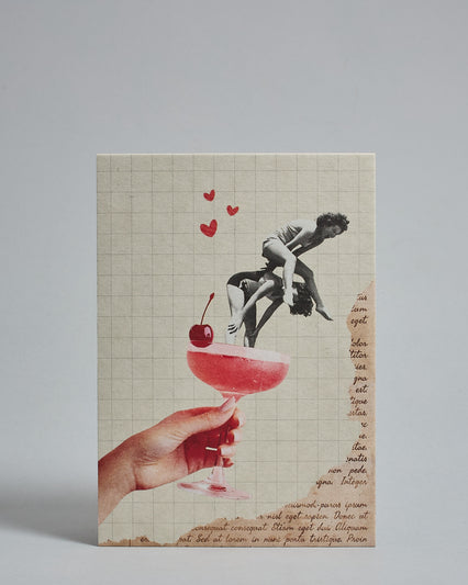 Die Vorderseite der Postkarte im Hochformat vor hellgrauem Hintergrund. Die Postkarte ist im Retro-Stil, gedruckt auf Holzschiffpappe. Im Vordergrund sind zwei Frauen in Graustufen die in einem Cocktail in Farbe springen. Im Hintergrund ist ein kariertes Papier und ein Ausschnitt aus einem alten Buch.