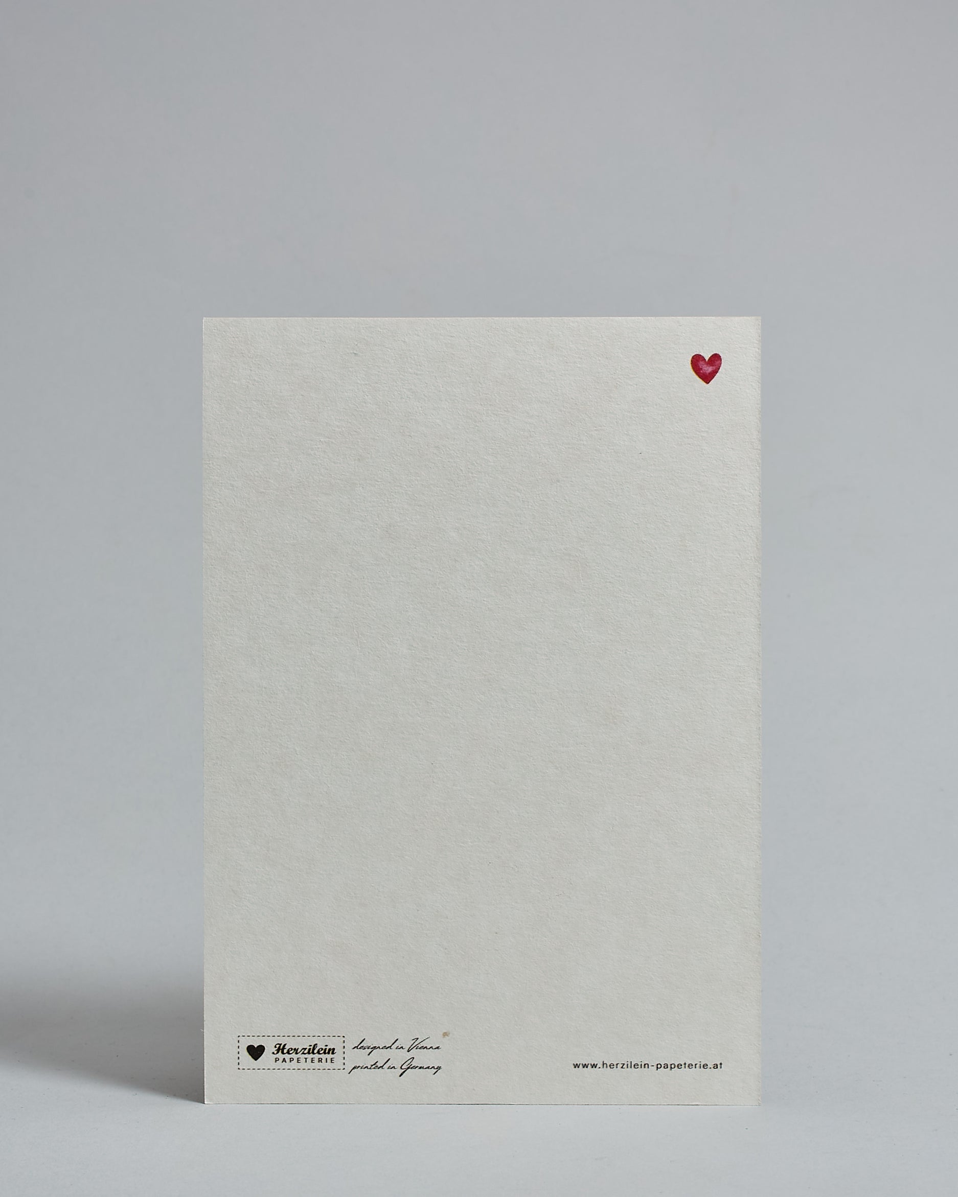 Die Rückseite der Postkarte im Hochformat vor hellgrauem Hintergrund. Rechts oben ist, in kleiner Ausführung, ein rotes Herz zu sehen. 
Unten sind klein das Logo Herzilein-Papeterie, der Text: "designed in VIenna, printed in Germany" und "www.herzilein-papeterie.at" zu sehen. 
Der Großteil ist unbedruckt die Struktur der hochwertigen Holzschliffpappe ist gut zu sehen. Hier ist viel Platz zum Schreiben.