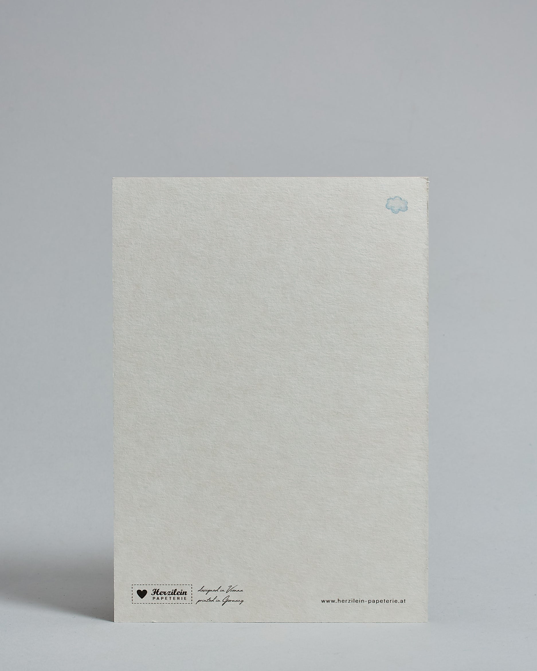 Die Rückseite der Postkarte im Hochformat vor hellgrauem Hintergrund. Rechts oben ist, in kleiner Ausführung, eine Wolke zu sehen. 
Unten sind klein das Logo Herzilein-Papeterie, der Text: "designed in VIenna, printed in Germany" und "www.herzilein-papeterie.at" zu sehen. 
Der Großteil ist unbedruckt die Struktur der hochwertigen Holzschliffpappe ist gut zu sehen. Hier ist viel Platz zum Schreiben.