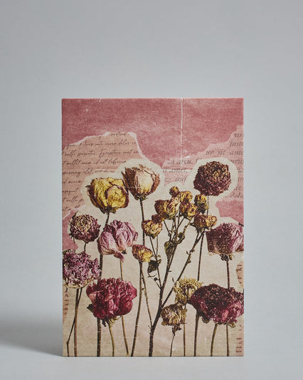 Die Vorderseite der Postkarte im Hochformat vor hellgrauem Hintergrund. Die Postkarte ist im Retro-Stil, gedruckt auf Holzschiffpappe und zeigt gemalt aussehende getrocknete Blumen im Vordergrund. Diese sehen aus als wäre sie aus einer Zeitschrift aufgeschnitten und auf den Hintergrund geklebt. Der Hintergrund besteht aus Stücken von Seiten aus alten Büchern auf einem lachsrotem Hintergrund.