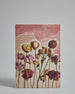 Die Vorderseite der Postkarte im Hochformat vor hellgrauem Hintergrund. Die Postkarte ist im Retro-Stil, gedruckt auf Holzschiffpappe und zeigt gemalt aussehende getrocknete Blumen im Vordergrund. Diese sehen aus als wäre sie aus einer Zeitschrift aufgeschnitten und auf den Hintergrund geklebt. Der Hintergrund besteht aus Stücken von Seiten aus alten Büchern auf einem lachsrotem Hintergrund.