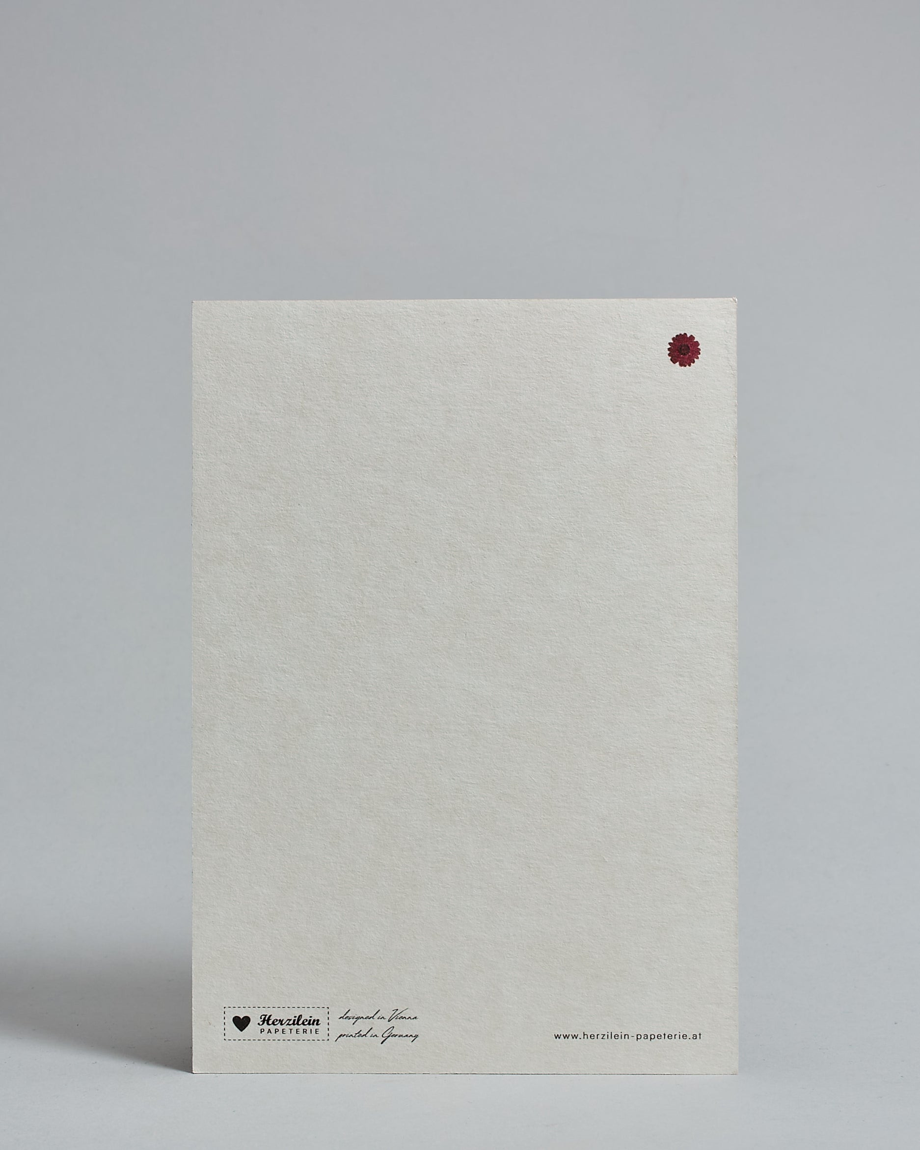 Die Rückseite der Postkarte im Hochformat vor hellgrauem Hintergrund. Rechts oben ist, in kleiner Ausführung, eine rote Blume zu sehen. 
Unten sind klein das Logo Herzilein-Papeterie, der Text: "designed in VIenna, printed in Germany" und "www.herzilein-papeterie.at" zu sehen. 
Der Großteil ist unbedruckt die Struktur der hochwertigen Holzschliffpappe ist gut zu sehen. Hier ist viel Platz zum Schreiben.
