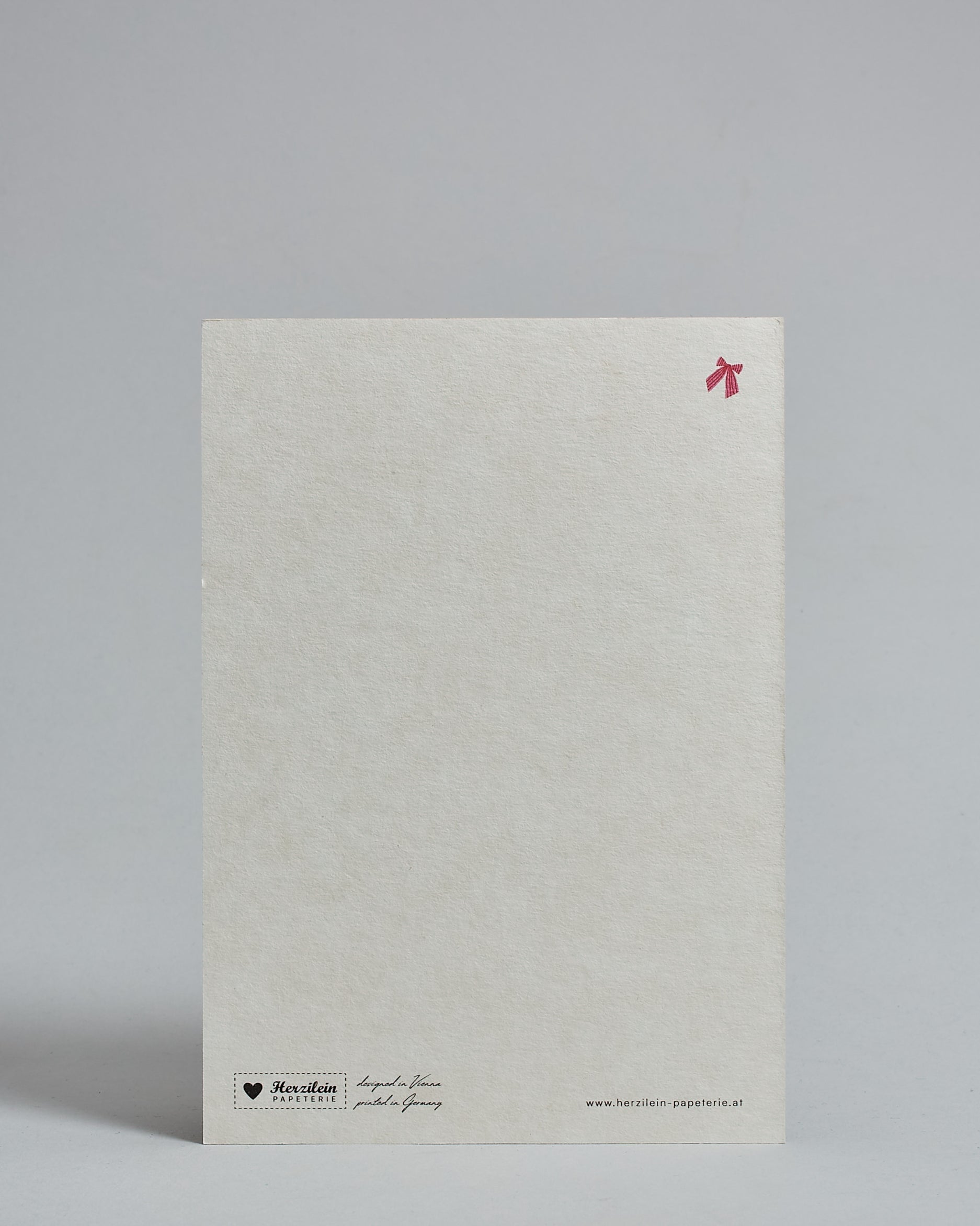 Die Rückseite der Postkarte im Hochformat vor hellgrauem Hintergrund. Rechts oben ist, in kleiner Ausführung, eine rote Masche zu sehen. 
Unten sind klein das Logo Herzilein-Papeterie, der Text: "designed in VIenna, printed in Germany" und "www.herzilein-papeterie.at" zu sehen. 
Der Großteil ist unbedruckt die Struktur der hochwertigen Holzschliffpappe ist gut zu sehen. Hier ist viel Platz zum Schreiben.