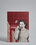Die Vorderseite der Postkarte im Hochformat vor hellgrauem Hintergrund. Die Postkarte ist im Retro-Stil, gedruckt auf Holzschiffpappe und zeigt eine Frau am Telefon in Graustufen vor einer klassisch britischen Telefonzelle. Der Hintergrund ist kariert dunkelrot und besteht zu einem Teil aus einer Seite aus einem alten Buch.