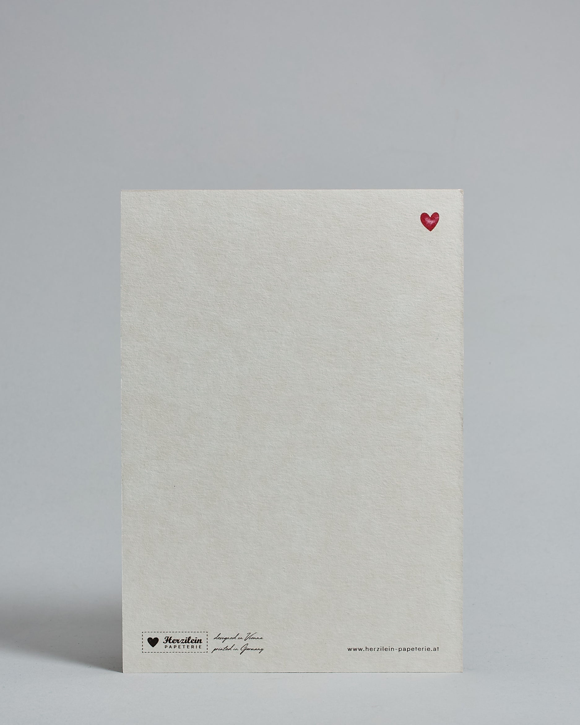 Die Rückseite der Postkarte im Hochformat vor hellgrauem Hintergrund. Rechts oben ist, in kleiner Ausführung, ein rotes Herz zu sehen. 
Unten sind klein das Logo Herzilein-Papeterie, der Text: "designed in VIenna, printed in Germany" und "www.herzilein-papeterie.at" zu sehen. 
Der Großteil ist unbedruckt die Struktur der hochwertigen Holzschliffpappe ist gut zu sehen. Hier ist viel Platz zum Schreiben.