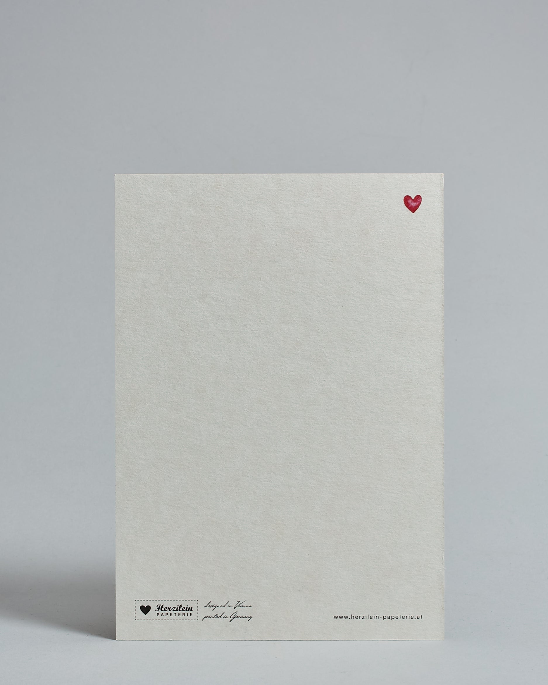 Die Rückseite der Postkarte im Hochformat vor hellgrauem Hintergrund. Rechts oben ist, in kleiner Ausführung, ein rotes Herz zu sehen. 
Unten sind klein das Logo Herzilein-Papeterie, der Text: "designed in VIenna, printed in Germany" und "www.herzilein-papeterie.at" zu sehen. 
Der Großteil ist unbedruckt die Struktur der hochwertigen Holzschliffpappe ist gut zu sehen. Hier ist viel Platz zum Schreiben.