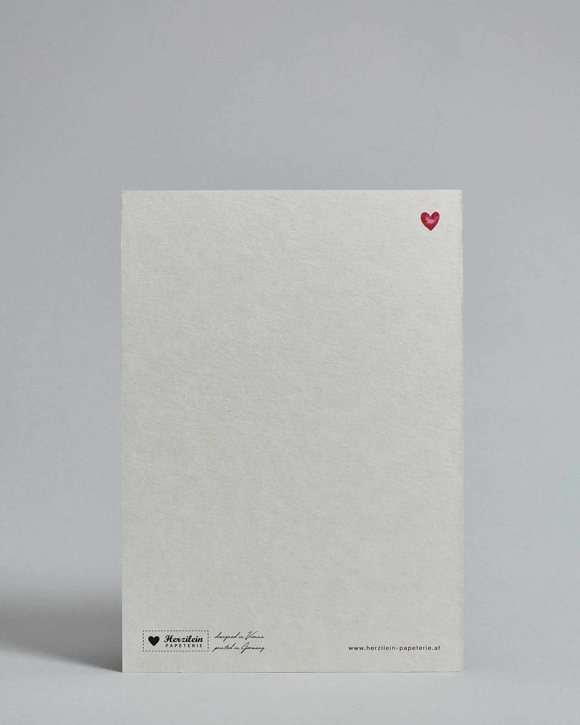 Die Rückseite der Postkarte im Hochformat vor hellgrauem Hintergrund. Rechts oben ist, in kleiner Ausführung, ein rotes Herz zu sehen. 
Unten sind klein das Logo Herzilein-Papeterie, der Text: "designed in VIenna, printed in Germany" und "www.herzilein-papeterie.at" zu sehen. 
Der Großteil ist unbedruckt die Struktur der hochwertigen Holzschliffpappe ist gut zu sehen. Hier ist viel Platz zum Schreiben.