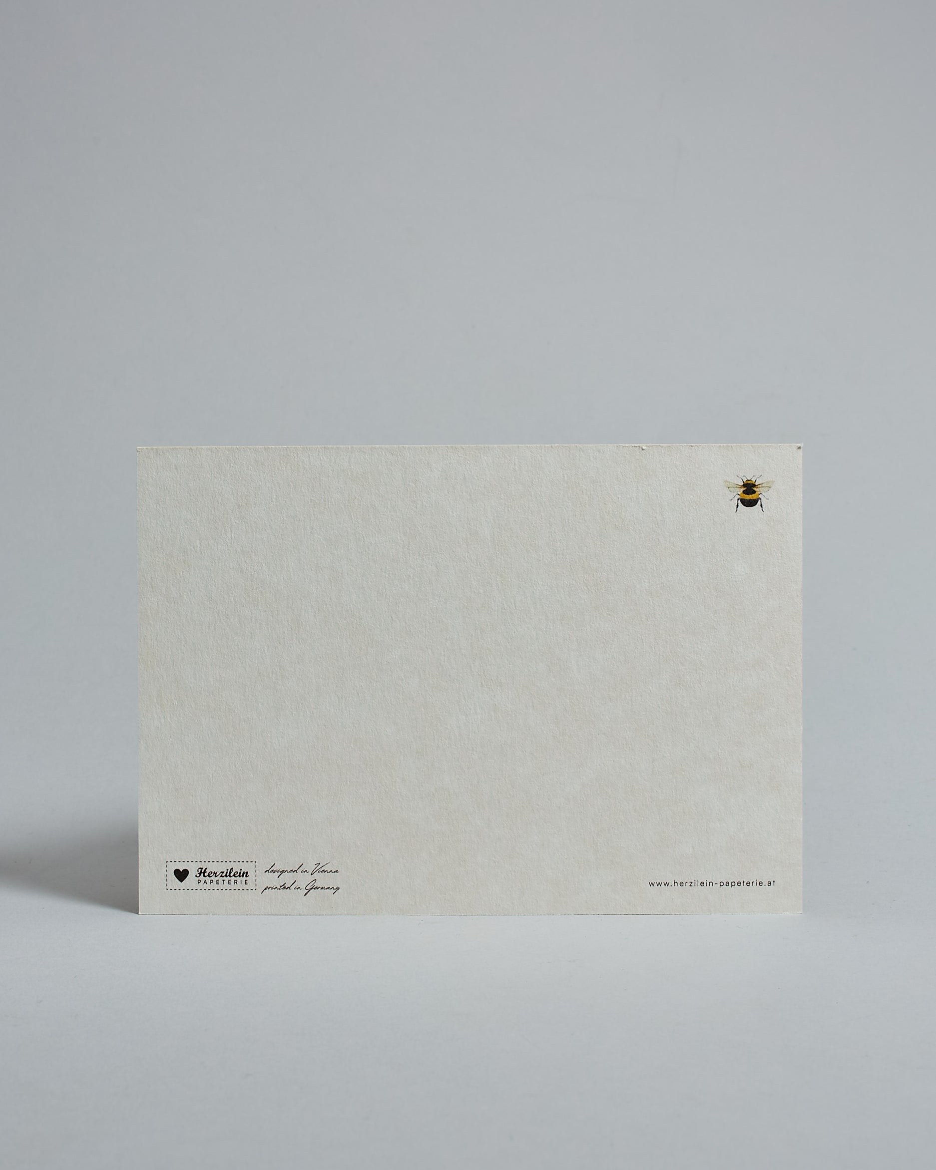 Die Rückseite der Postkarte im Querformat vor hellgrauem Hintergrund. Rechts oben ist, in kleiner Ausführung, die gleiche Biene wie auf der Vorderseite zu sehen. 
Unten sind klein das Logo Herzilein-Papeterie, der Text: "designed in VIenna, printed in Germany" und "www.herzilein-papeterie.at" zu sehen. 
Der Großteil ist unbedruckt die Struktur der hochwertigen Holzschliffpappe ist gut zu sehen. Hier ist viel Platz zum Schreiben.