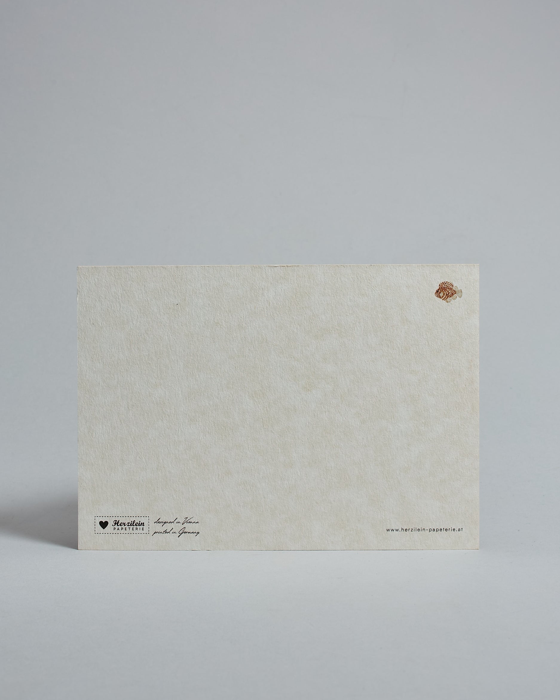 Die Rückseite der Postkarte im Querformat vor hellgrauem Hintergrund. Rechts oben ist, in kleiner Ausführung, der gleiche Feuerfisch wie auf der Vorderseite zu sehen. 
Unten sind klein das Logo Herzilein-Papeterie, der Text: "designed in VIenna, printed in Germany" und "www.herzilein-papeterie.at" zu sehen. 
Der Großteil ist unbedruckt die Struktur der hochwertigen Holzschliffpappe ist gut zu sehen. Hier ist viel Platz zum Schreiben.