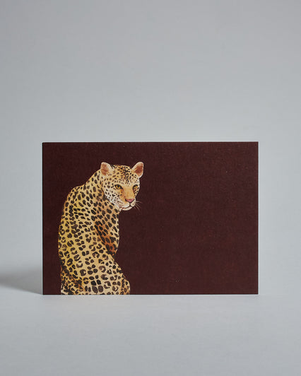 Die Vorderseite der Postkarte im Querformat vor hellgrauem Hintergrund. Die Postkarte zeigt einen geamlten Leopoarden auf braunrotem Hintergrund, gedruckt auf Holzschliffpappe.