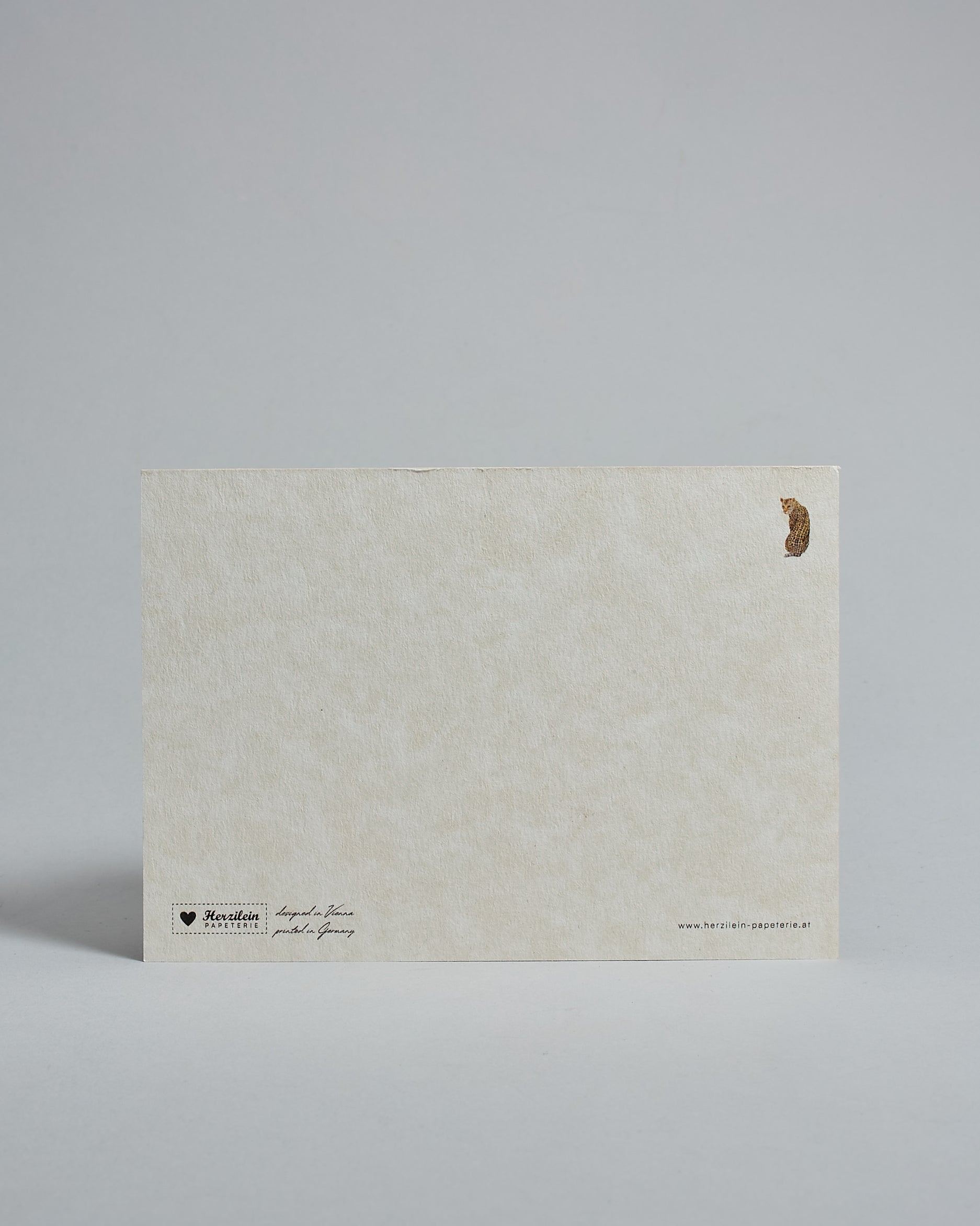 Die Rückseite der Postkarte im Querformat vor hellgrauem Hintergrund. Rechts oben ist, in kleiner Ausführung, der gleiche Leopard wie auf der Vorderseite zu sehen. 
Unten sind klein das Logo Herzilein-Papeterie, der Text: "designed in VIenna, printed in Germany" und "www.herzilein-papeterie.at" zu sehen. 
Der Großteil ist unbedruckt die Struktur der hochwertigen Holzschliffpappe ist gut zu sehen. Hier ist viel Platz zum Schreiben.