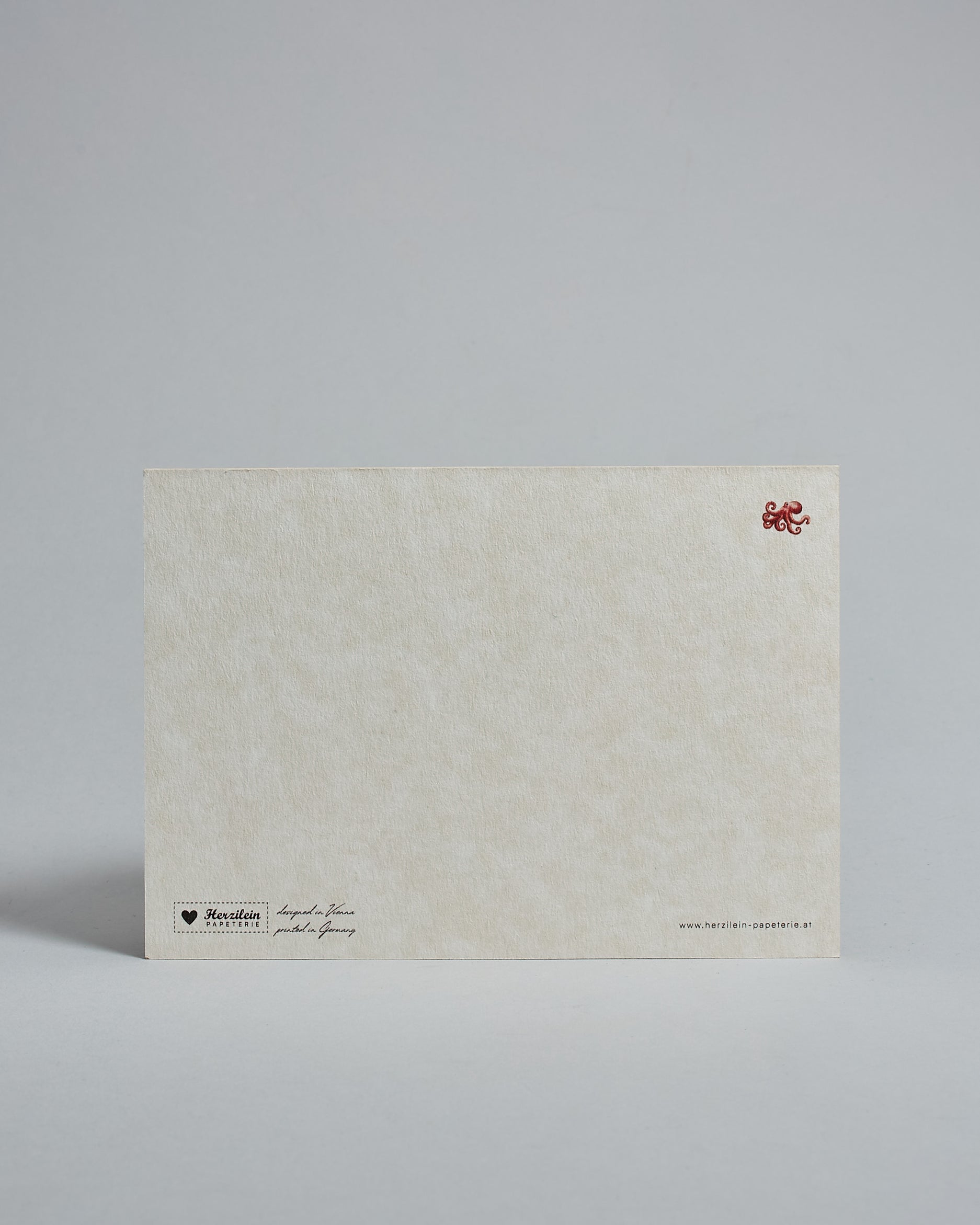 Die Rückseite der Postkarte im Querformat vor hellgrauem Hintergrund. Rechts oben ist, in kleiner Ausführung, der gleiche Tintenfisch wie auf der Vorderseite zu sehen. 
Unten sind klein das Logo Herzilein-Papeterie, der Text: "designed in VIenna, printed in Germany" und "www.herzilein-papeterie.at" zu sehen. 
Der Großteil ist unbedruckt die Struktur der hochwertigen Holzschliffpappe ist gut zu sehen. Hier ist viel Platz zum Schreiben.