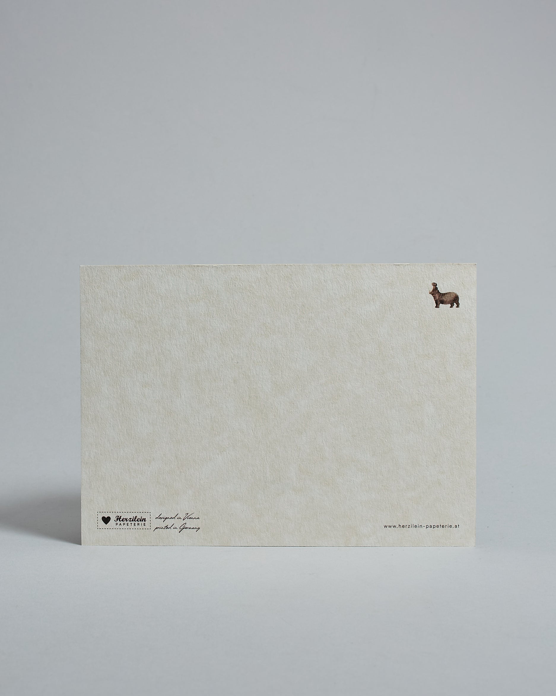 Die Rückseite der Postkarte im Querformat vor hellgrauem Hintergrund. Rechts oben ist, in kleiner Ausführung, das gleiche Nilpferd wie auf der Vorderseite zu sehen. 
Unten sind klein das Logo Herzilein-Papeterie, der Text: "designed in VIenna, printed in Germany" und "www.herzilein-papeterie.at" zu sehen. 
Der Großteil ist unbedruckt die Struktur der hochwertigen Holzschliffpappe ist gut zu sehen. Hier ist viel Platz zum Schreiben.