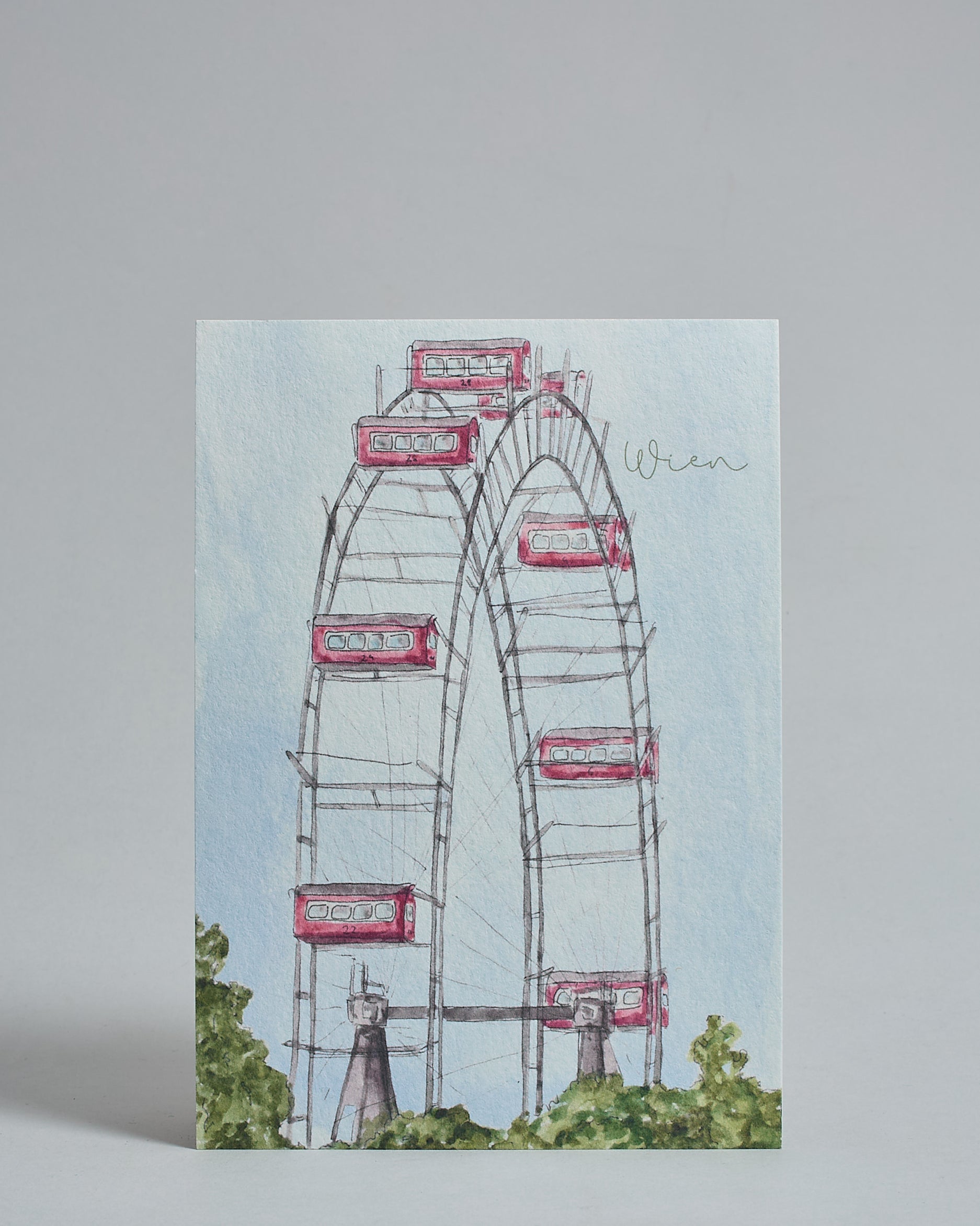 Die Vorderseite der Postkarte im Hochformat, gedruckt auf hochwertigem Papier. Diese zeigt die das Wiener Riesenrad vor blauem Himmel gemalt von einer Wiener Künstlerin. Rechts oben ist "Wien" in geschwunger, dezenter Schrift zu lesen. Im unteren Teil sieht man einige grüne Baumwipfel.