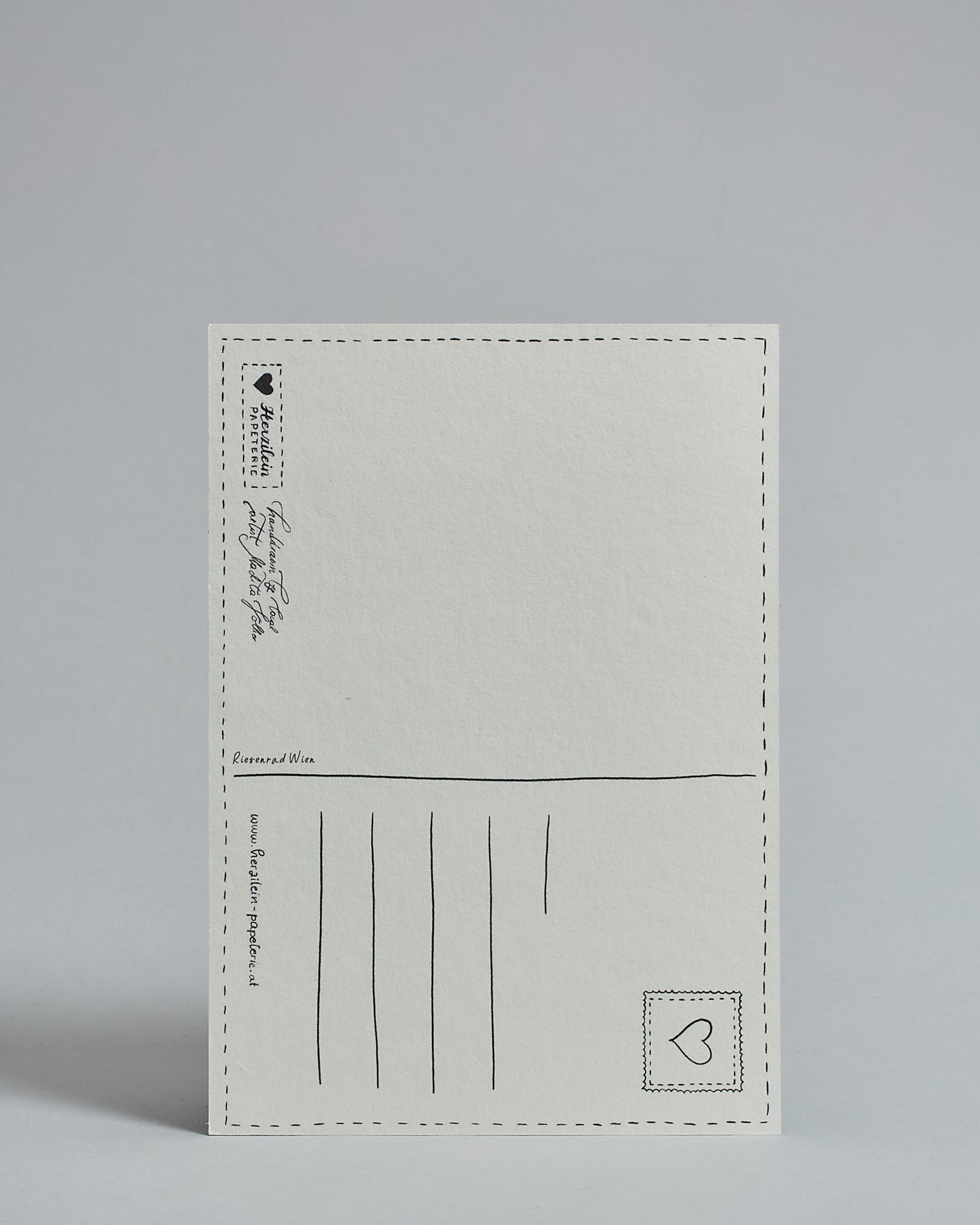 Die Rückseite der Postkarte im Hochfornat vor hellgrauem Hintergrund. 
Ein handgezeichnetes, Postkarten-typisches Layout mit Adressfeld auf der rechten Seite und Textfeld auf der linken Seite ist zu sehen, dazwischen eine Trennlinie. Auf der Trennlinie steht der auf der Vorderseite dargestellte Ort.  Rechts oben ist eine stilisiere Briefmarke mit Herz-Motiv zu sehen. Links unten steht das Logo Herzilein-Papeterie und daneben der Text: "handdrawn by local artist Madita Völker".