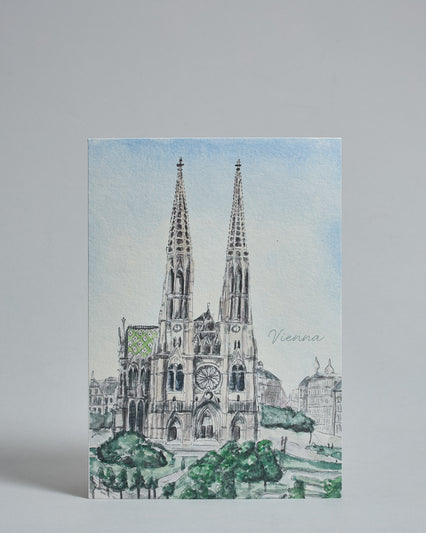 Die Vorderseite der Postkarte im Hochformat, gedruckt auf hochwertigem Papier. Diese zeigt die Wiener Votivkirche vor blauem Himmel gemalt von einer Wiener Künstlerin. Im unteren Teil der Karte ist ein Teil des Votivkparks zu sehen. rechts neben der Kirche auf mittlerer Höhe ist "Vienna" in dezenter, geschwungener Schrift zu lesen.