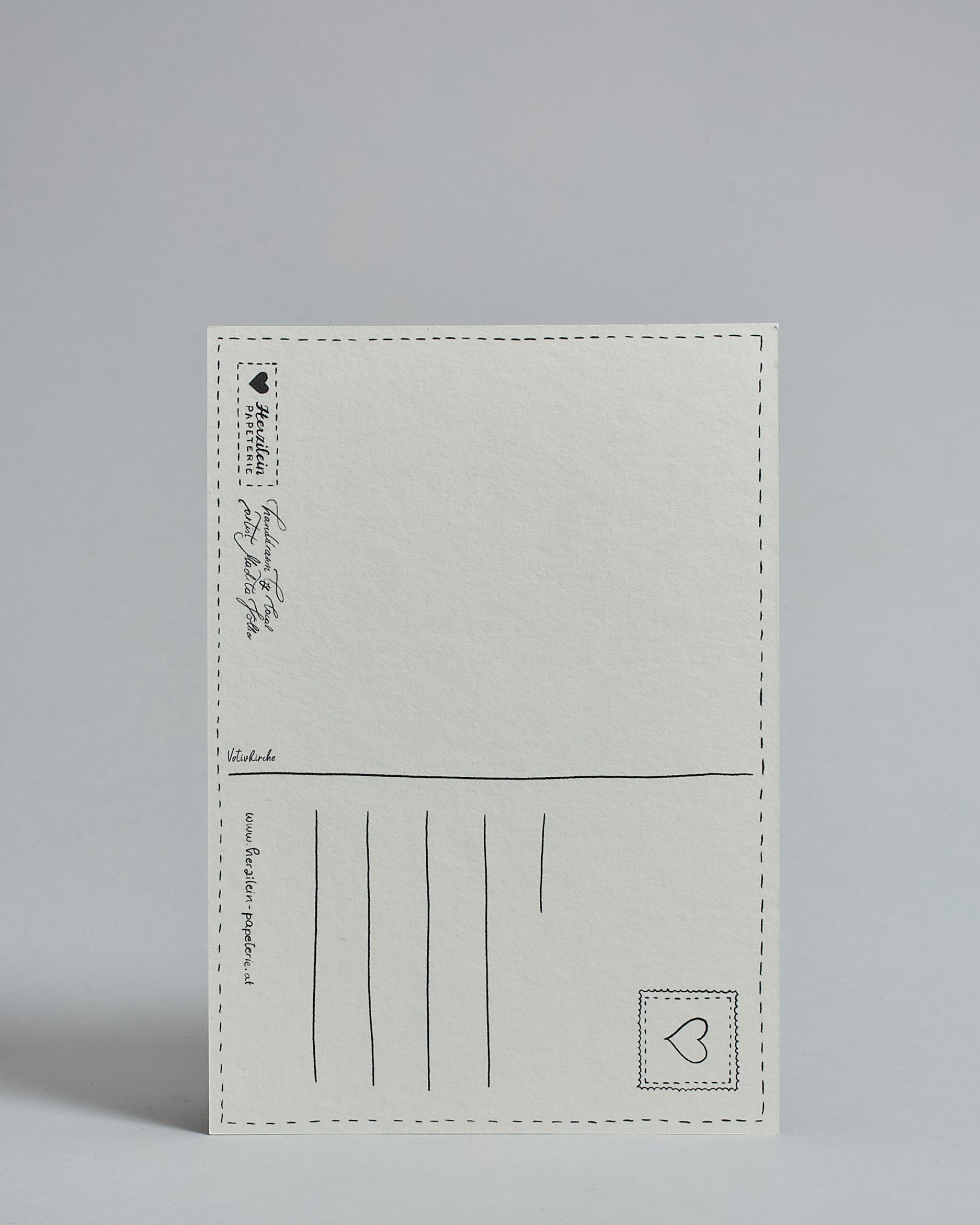 Die Rückseite der Postkarte im Hochfornat vor hellgrauem Hintergrund. 
Ein handgezeichnetes, Postkarten-typisches Layout mit Adressfeld auf der rechten Seite und Textfeld auf der linken Seite ist zu sehen, dazwischen eine Trennlinie. Auf der Trennlinie steht der auf der Vorderseite dargestellte Ort.  Rechts oben ist eine stilisiere Briefmarke mit Herz-Motiv zu sehen. Links unten steht das Logo Herzilein-Papeterie und daneben der Text: "handdrawn by local artist Madita Völker".