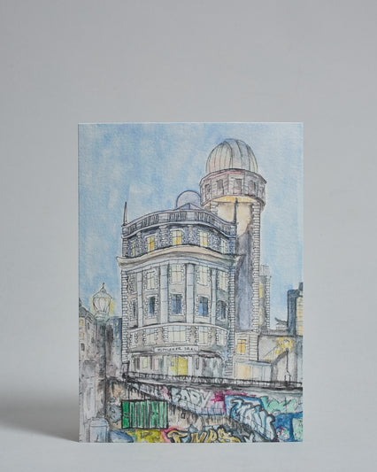 Die Vorderseite der Postkarte im Hochformat, gedruckt auf hochwertigem Papier. Diese zeigt die Wiener Urania vor blauem Himmelgemalt von einer Wiener Künstlerin. Im unteren Teil sind bunte Graffitis vom Donaukanal sichtbar.