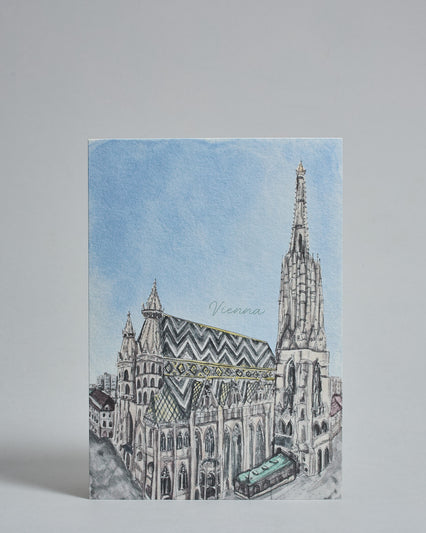 Die Vorderseite der Postkarte im Hochformat, gedruckt auf hochwertigem Papier. Diese zeigt den Wiener Stephansdom vor blauem Himmel gemalt von einer Wiener Künstlerin. Über dem Gibel ist der Text "Vienna" in dezenter, geschwunger Schrift zu lesen.
