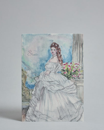 Die Vorderseite der Postkarte im Hochformat, gedruckt auf hochwertigem Papier. Diese zeigt die Kaiserin Sisi gemalt von einer Wiener Künstlerin. Auf der linken Seite ist "Sisi" in dezenter, geschwungener Schrift zu lesen.