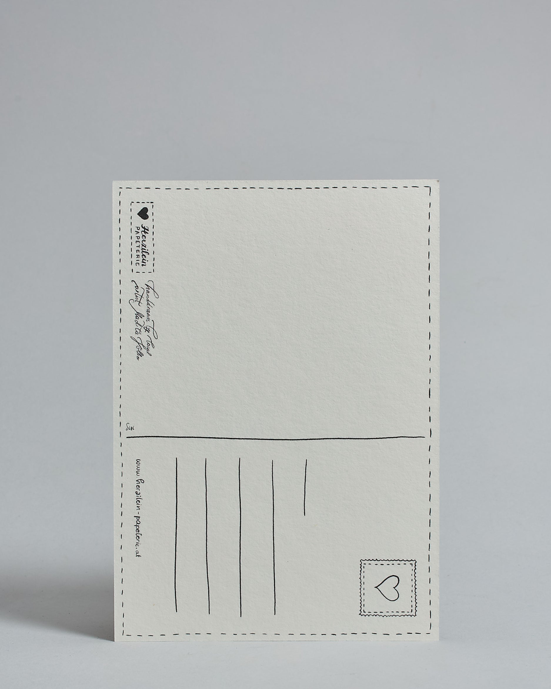 Die Rückseite der Postkarte im Hochfornat vor hellgrauem Hintergrund. 
Ein handgezeichnetes, Postkarten-typisches Layout mit Adressfeld auf der rechten Seite und Textfeld auf der linken Seite ist zu sehen, dazwischen eine Trennlinie. Auf der Trennlinie steht der auf der Vorderseite dargestellte Ort.  Rechts oben ist eine stilisiere Briefmarke mit Herz-Motiv zu sehen. Links unten steht das Logo Herzilein-Papeterie und daneben der Text: "handdrawn by local artist Madita Völker".
