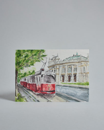 Die Vorderseite der Postkarte im Querformat, gedruckt auf hochwertigem Papier. Diese zeigt eine alte Wiener Straßenbahn vor dem Burgtheater gemalt von einer Wiener Künstlerin. Anstelle von Fahrziel und Liniennummer sind ein Herz und "Herzilein" auf der Straßenbahn zu lesen. Im linken Bildteil ist ein Baum, dessen grüne Krone teilweise über die Straßenbahn reicht.