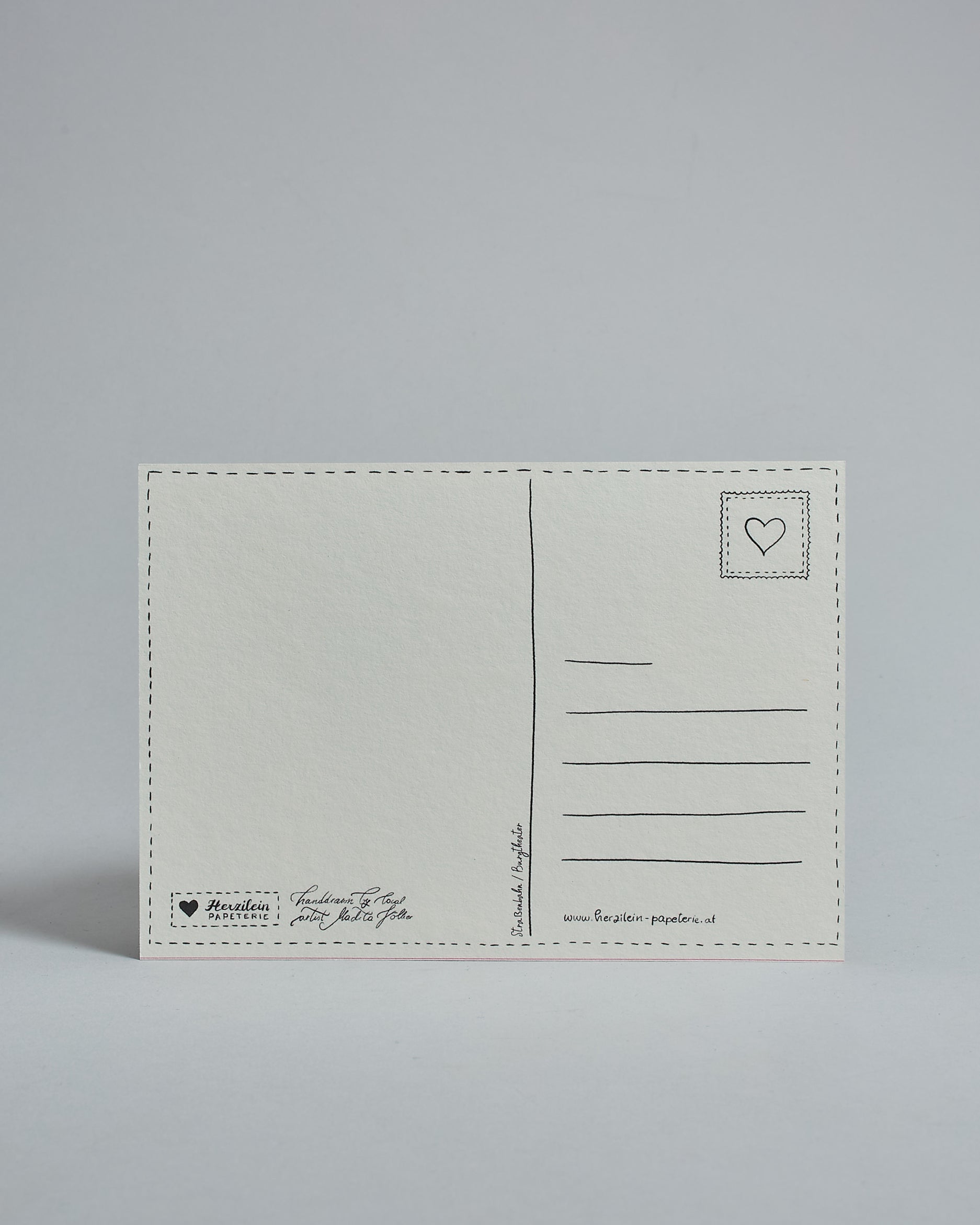 Die Rückseite der Postkarte im Querformat vor hellgrauem Hintergrund. 
Ein handgezeichnetes, Postkarten-typisches Layout mit Adressfeld auf der rechten Seite und Textfeld auf der linken Seite ist zu sehen, dazwischen eine Trennlinie. Auf der Trennlinie steht der auf der Vorderseite dargestellte Ort.  Rechts oben ist eine stilisiere Briefmarke mit Herz-Motiv zu sehen. Links unten steht das Logo Herzilein-Papeterie und daneben der Text: "handdrawn by local artist Madita Völker".
