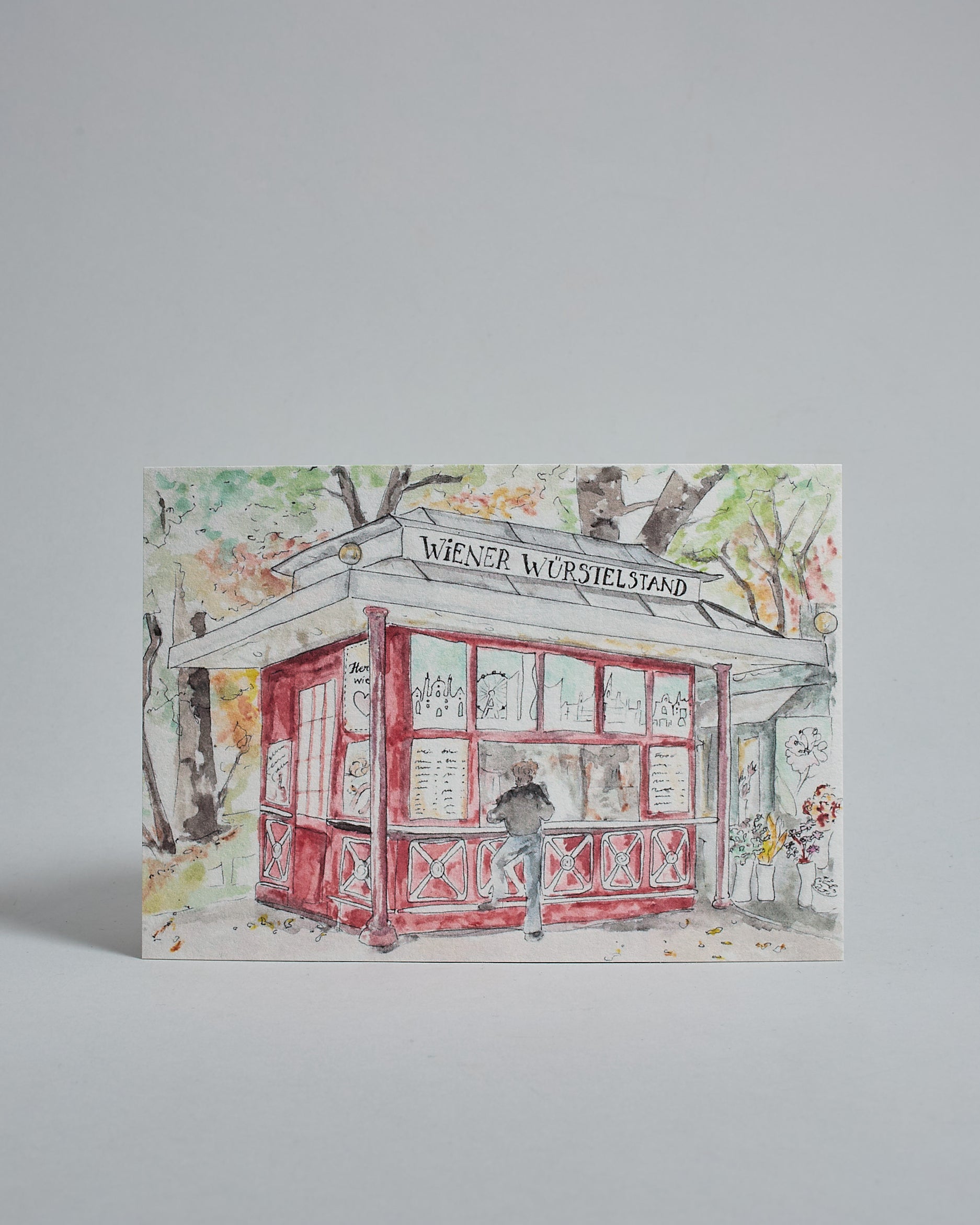 Die Vorderseite der Postkarte im Querformat, gedruckt auf hochwertigem Papier. Diese zeigt die einen Wiener Würstelstand vor dem Stadtpark gemalt von einer Wiener Künstlerin. Auf dem roten Stand ist ein Schild mit dem Aufdruck "Wiener Würstelstand" zu sehen. Im Hintergrund sind herbstliche Bäume des Stadtparks zu sehen.