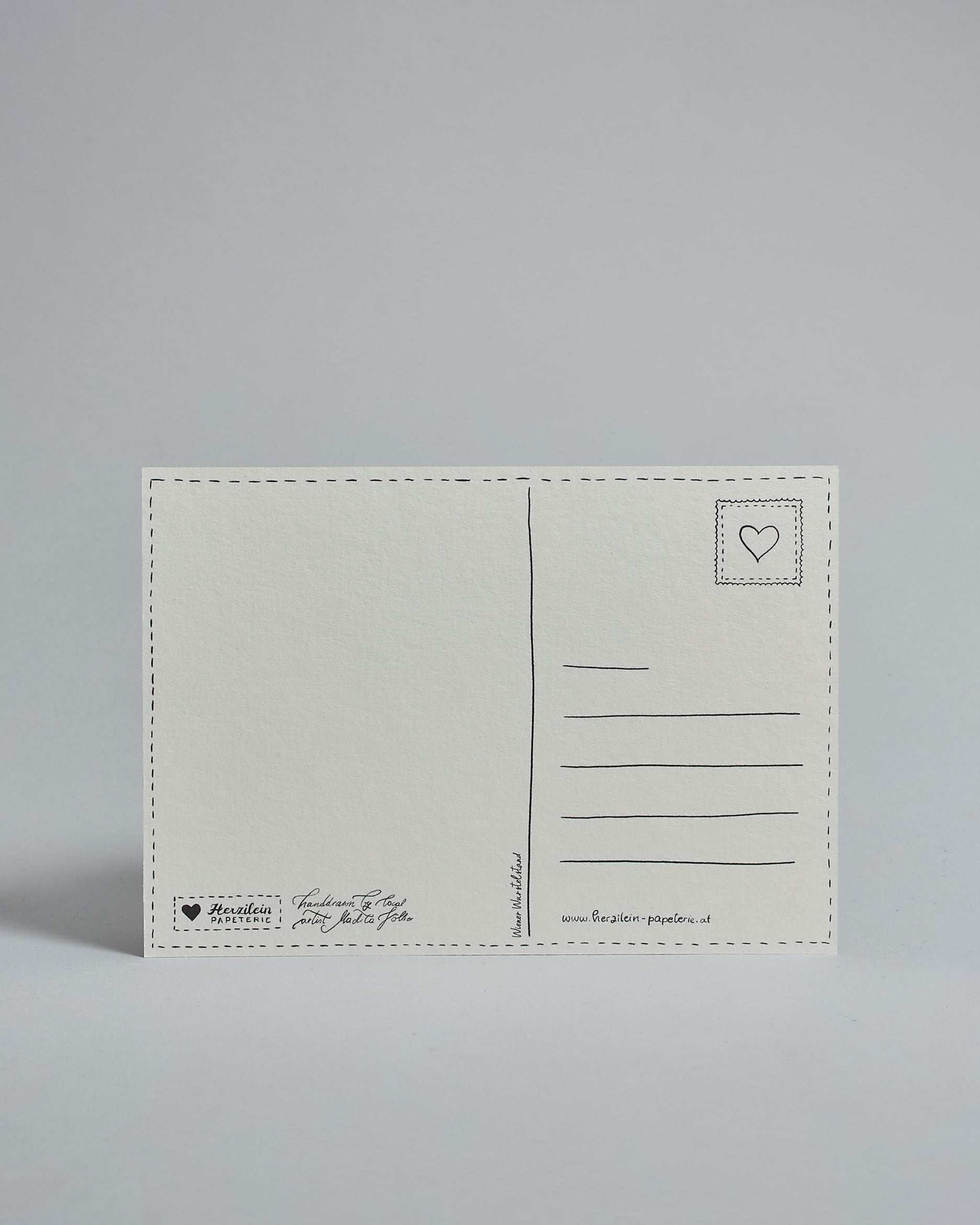 Die Rückseite der Postkarte im Querformat vor hellgrauem Hintergrund. 
Ein handgezeichnetes, Postkarten-typisches Layout mit Adressfeld auf der rechten Seite und Textfeld auf der linken Seite ist zu sehen, dazwischen eine Trennlinie. Auf der Trennlinie steht der auf der Vorderseite dargestellte Ort.  Rechts oben ist eine stilisiere Briefmarke mit Herz-Motiv zu sehen. Links unten steht das Logo Herzilein-Papeterie und daneben der Text: "handdrawn by local artist Madita Völker".