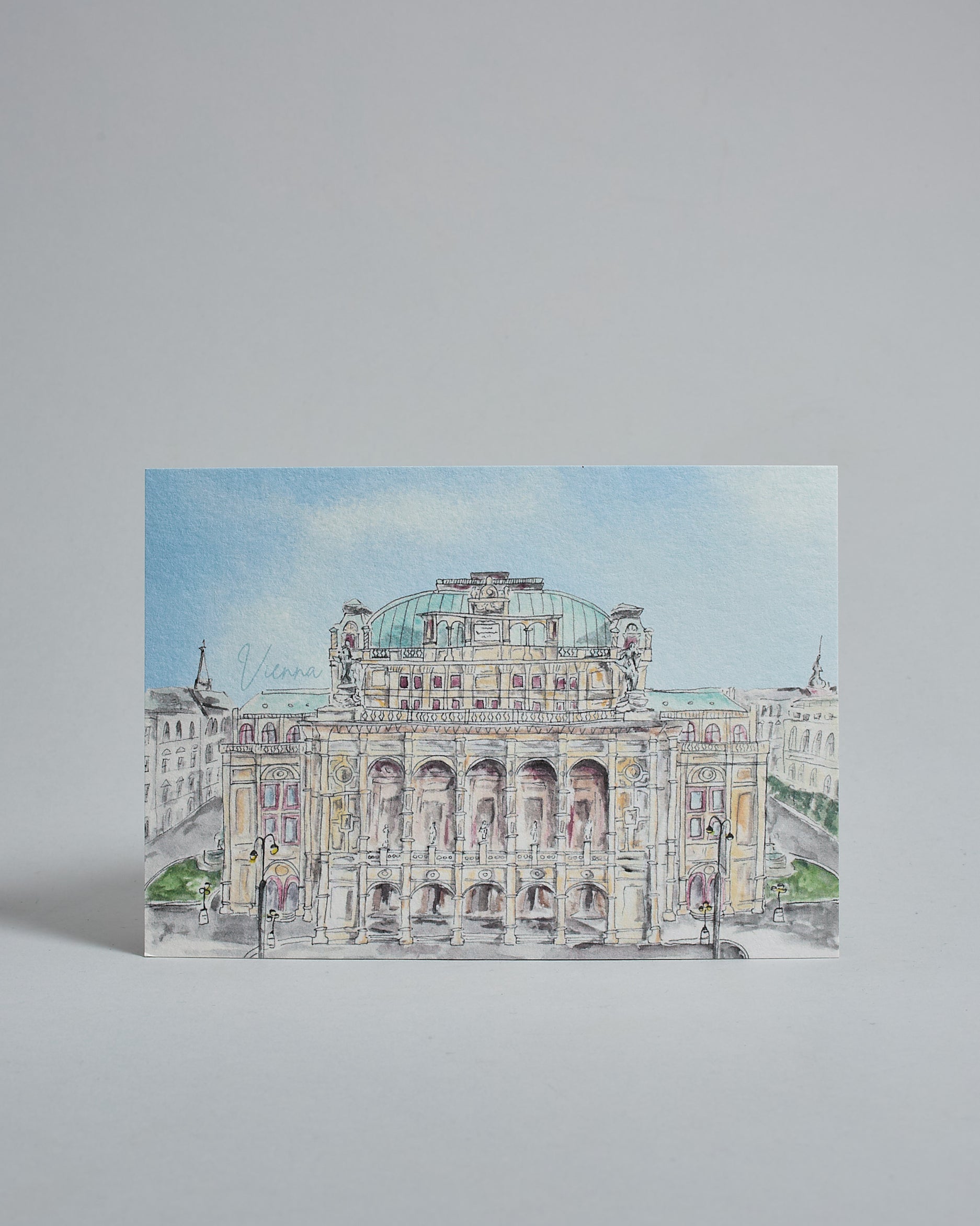 Die Vorderseite der Postkarte im Querformat, gedruckt auf hochwertigem Papier. Diese zeigt die Wiener Staatsoper vor blauem Himmel gemalt von einer Wiener Künstlerin. Links neben dem Gebäude ist der Schriftzug "Vienna" in dezenter, geschwungener Schrift zu lesen.
